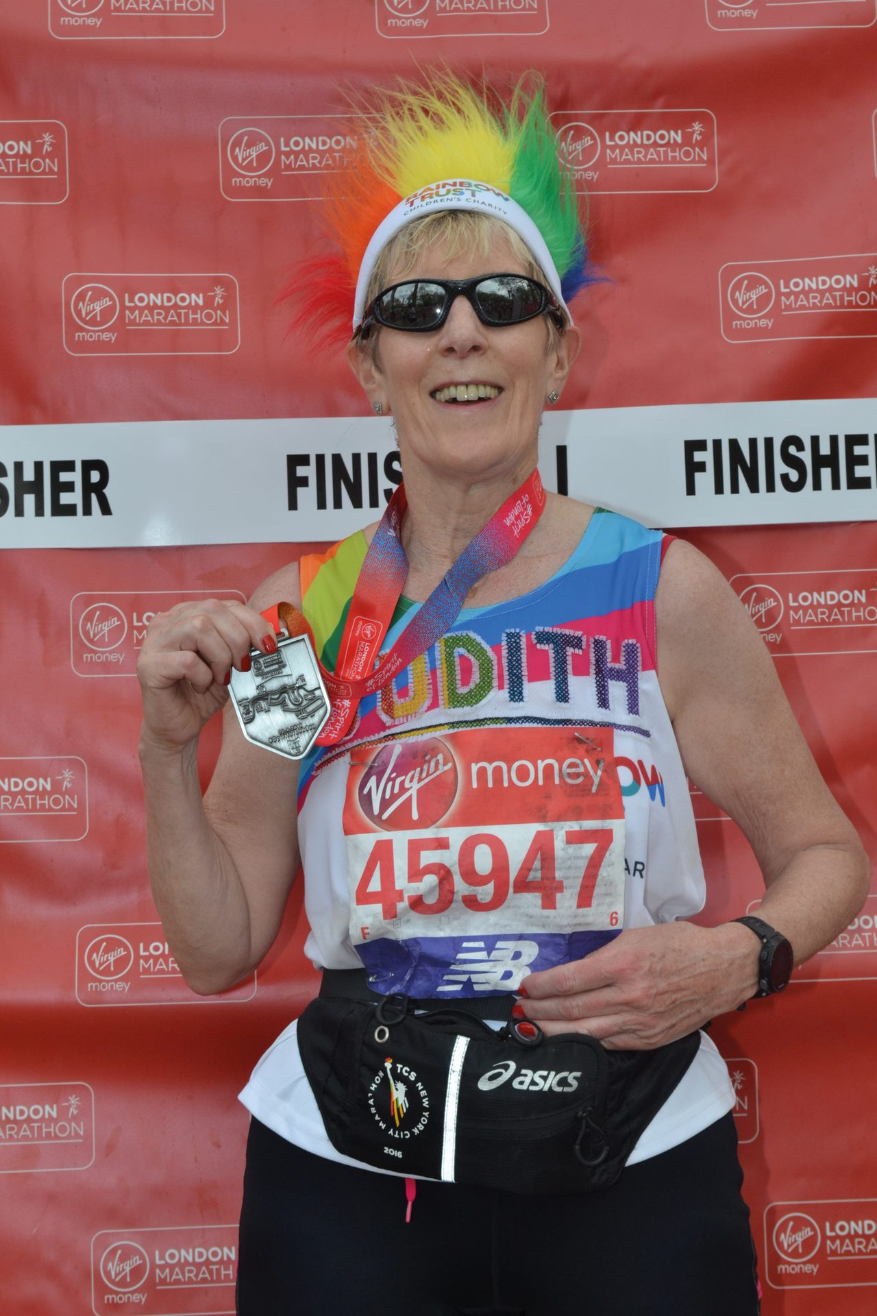 Woman in a rainbow wig and sunglasses, smiling, holding a medal after the London Marathon.