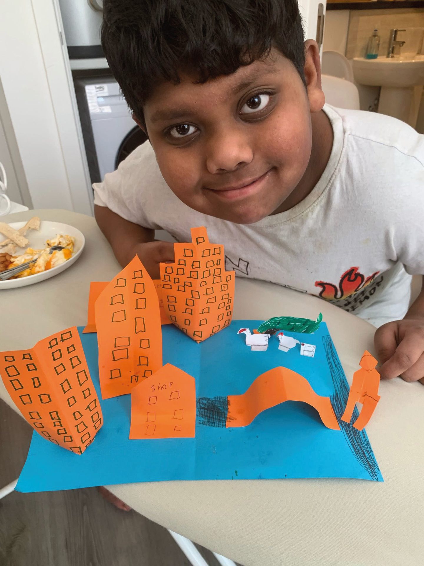 A smiling child proudly presents their colorful paper craft diorama with buildings and animals.