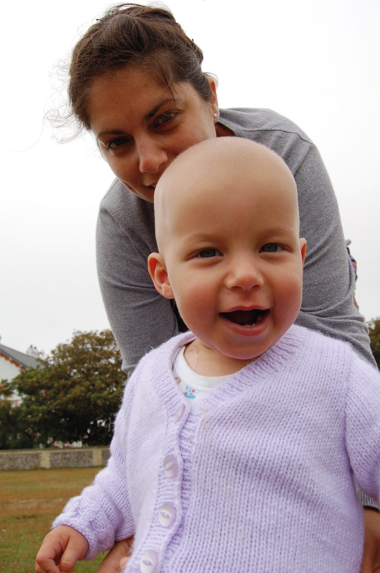 A mother smiles behind her baby, who is giggling in a lavender cardigan outdoors on a cloudy day.
