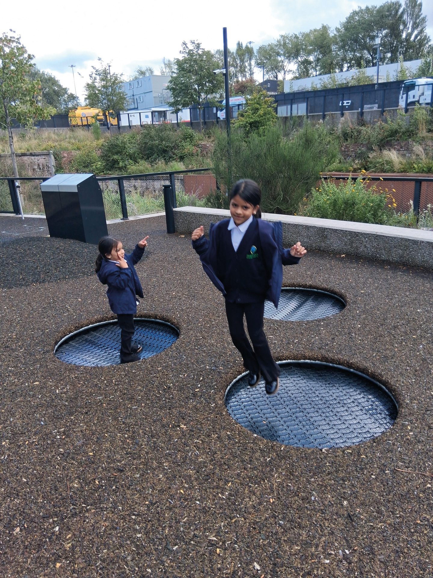 Two young girls in school uniforms playing on ground trampolines in an outdoor area.