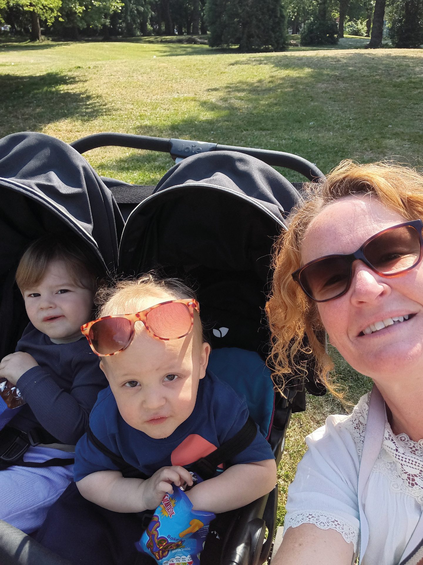 A woman takes a selfie with two babies in a double stroller in a sunny park. One baby wears sunglasses.