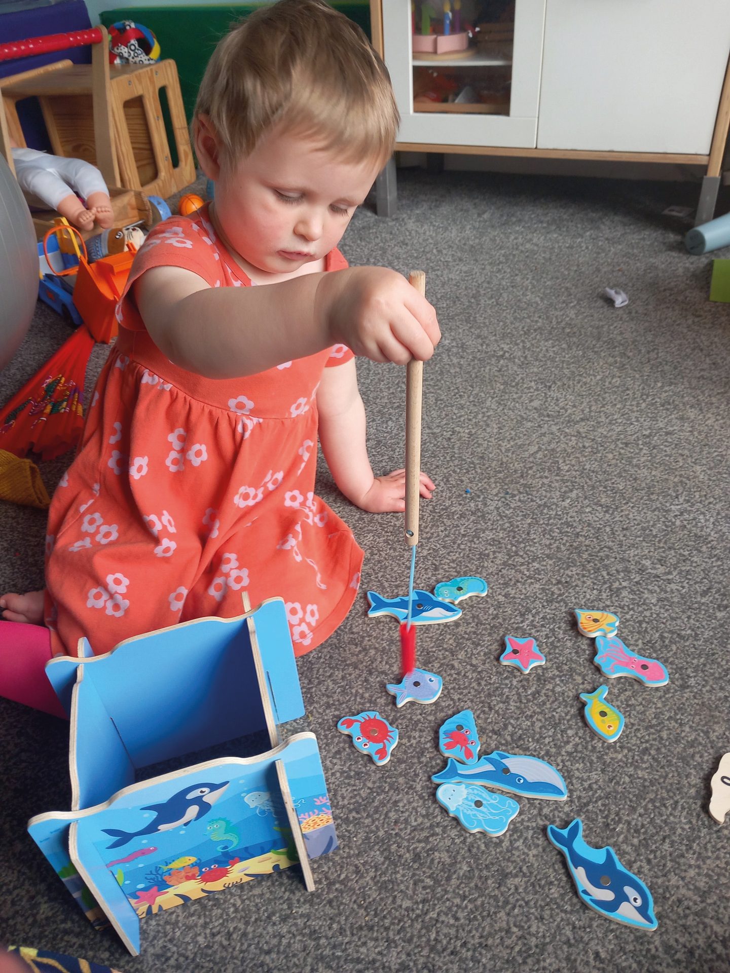 Toddler in orange dress playing magnetic fishing game on carpet, catching toy sea creatures.