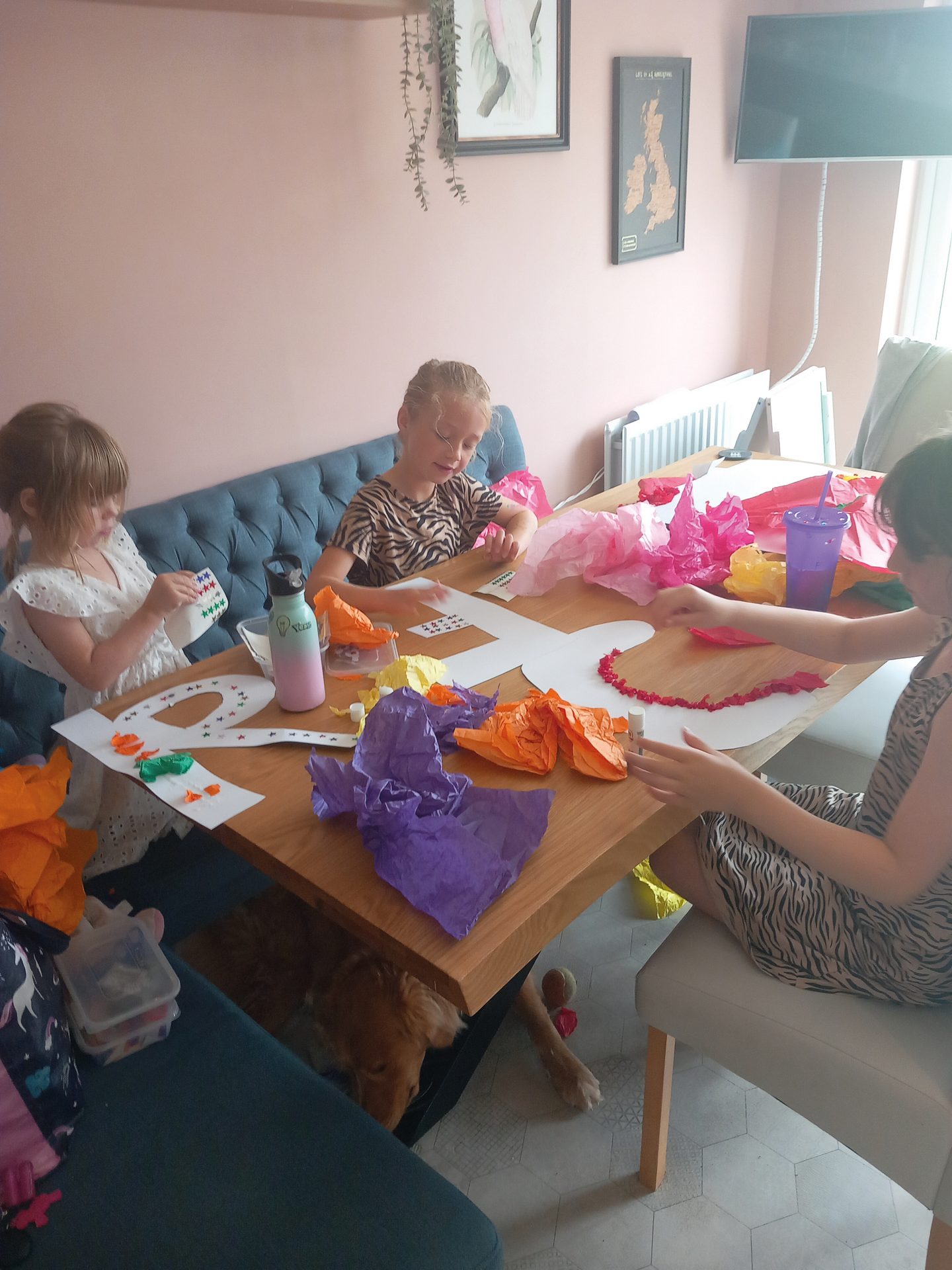 Three girls craft with colorful paper at a table; a dog rests underneath.