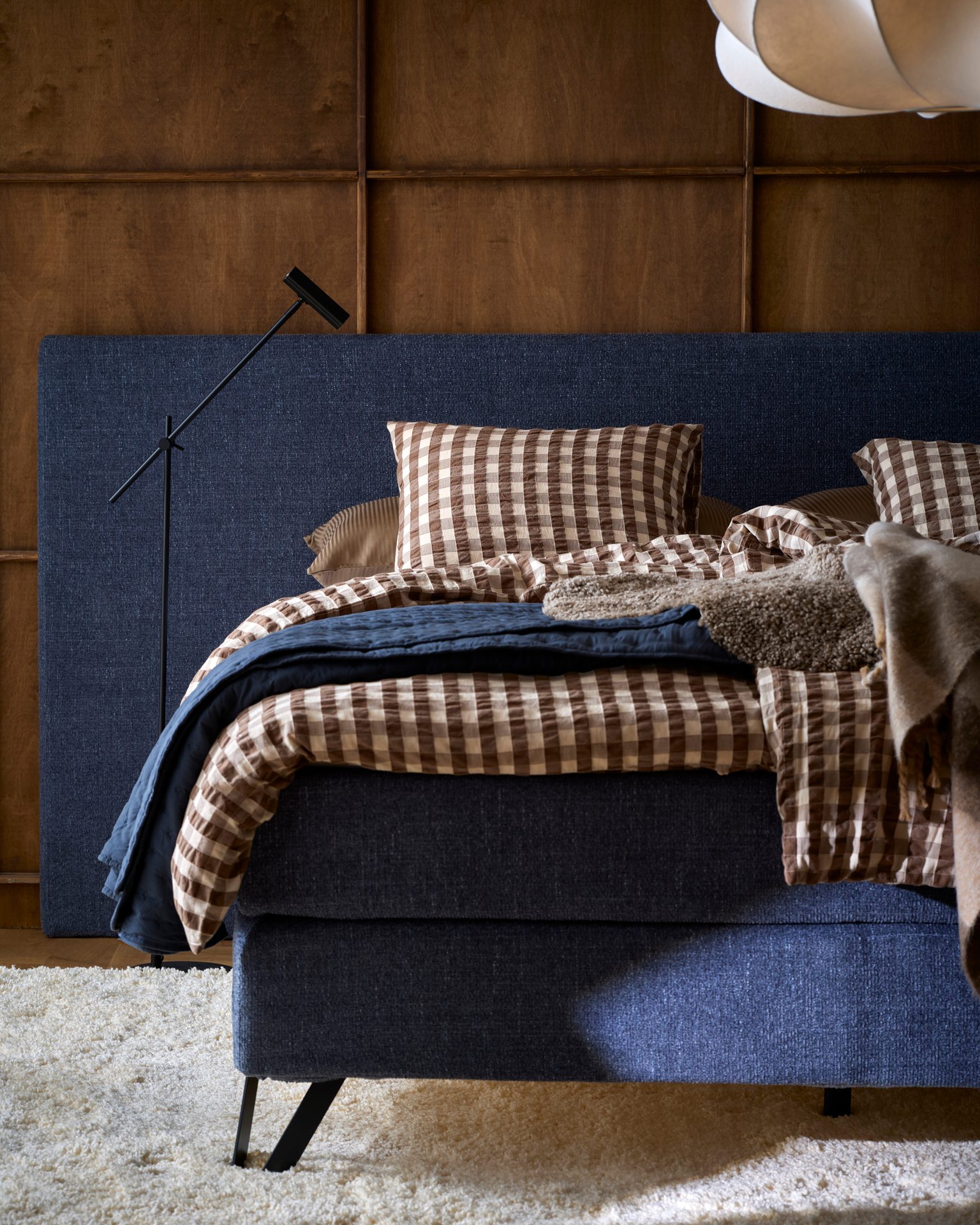 A cozy bedroom featuring a blue bed with a checked duvet, a floor lamp, and a wooden wall.