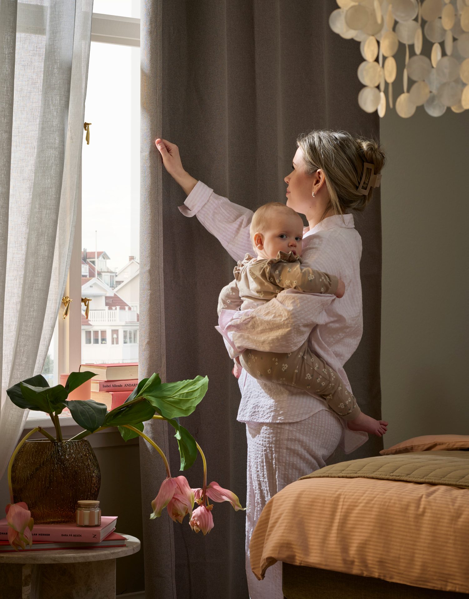 A mother in pajamas holds her baby while adjusting curtains and looking out a sunlit window in a bedroom.
