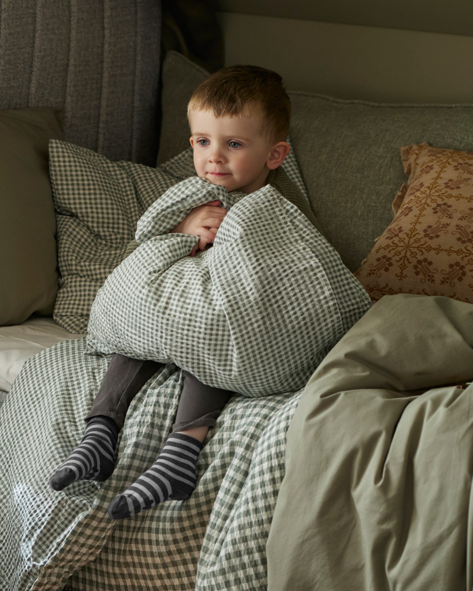 A cute little boy wrapped in a green and white checkered blanket, sitting on a bed.