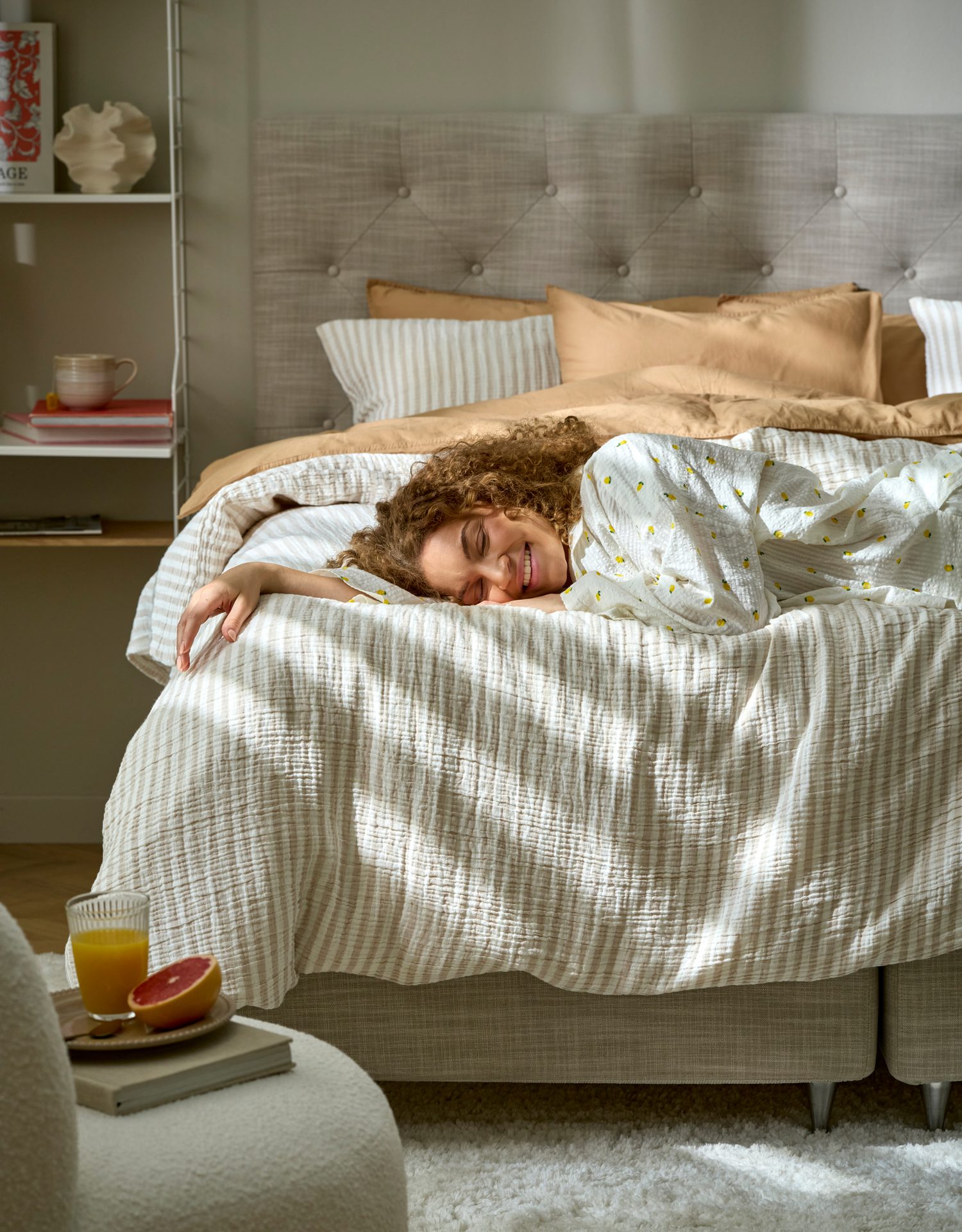 Woman with curly hair smiling and stretching in bed, with juice and grapefruit on a nearby table.