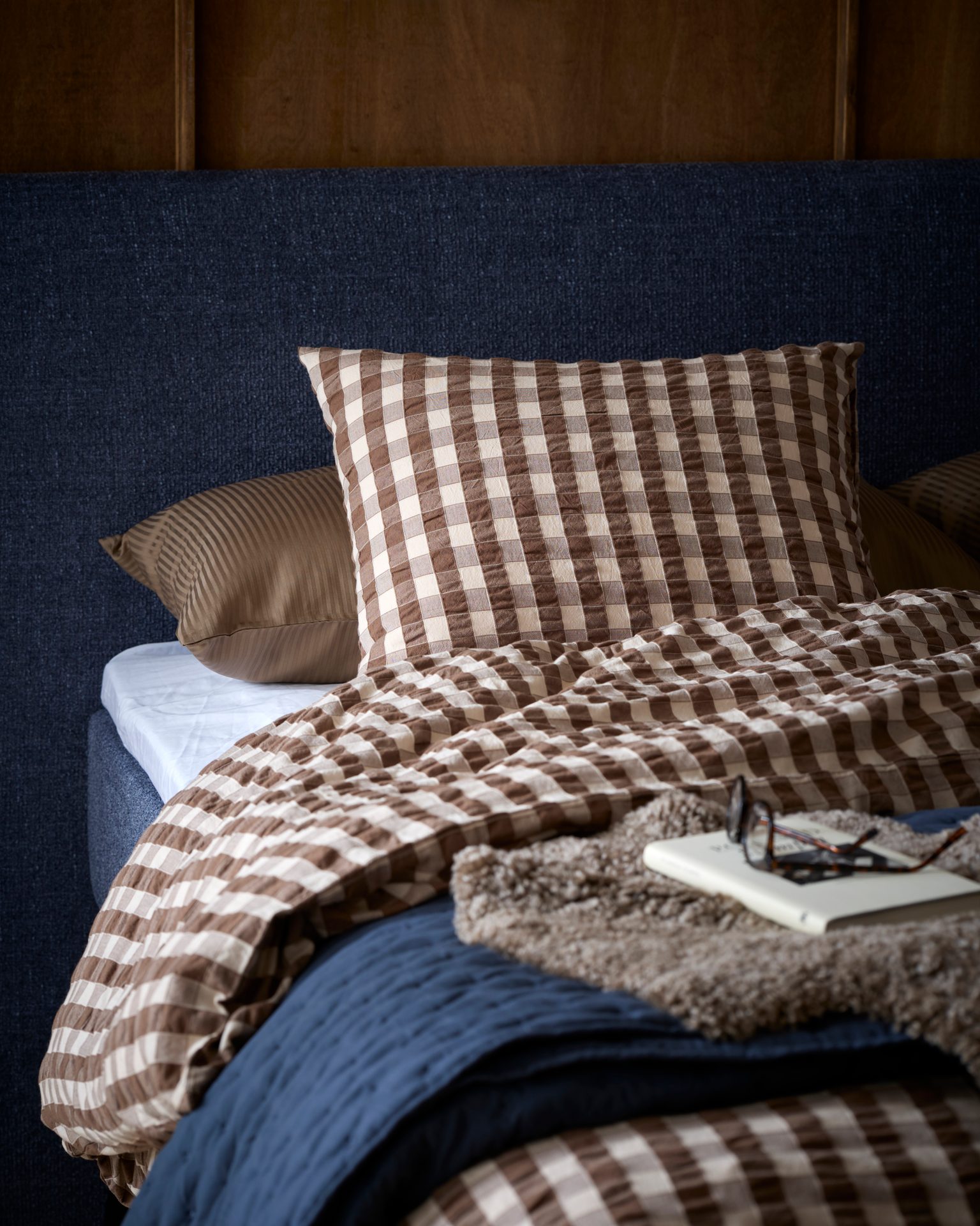 A cozy bed with a blue headboard, brown and white checkered duvet, striped pillow, blue blanket, book, and glasses.