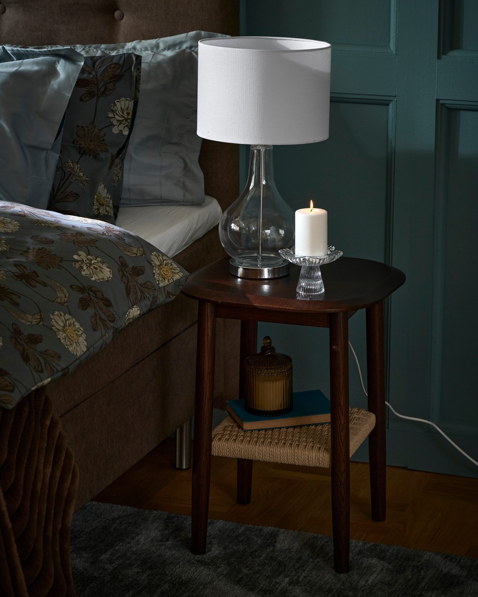 A cozy bedroom corner with a bed, a nightstand, a table lamp, and a lit candle, creating a warm ambiance.