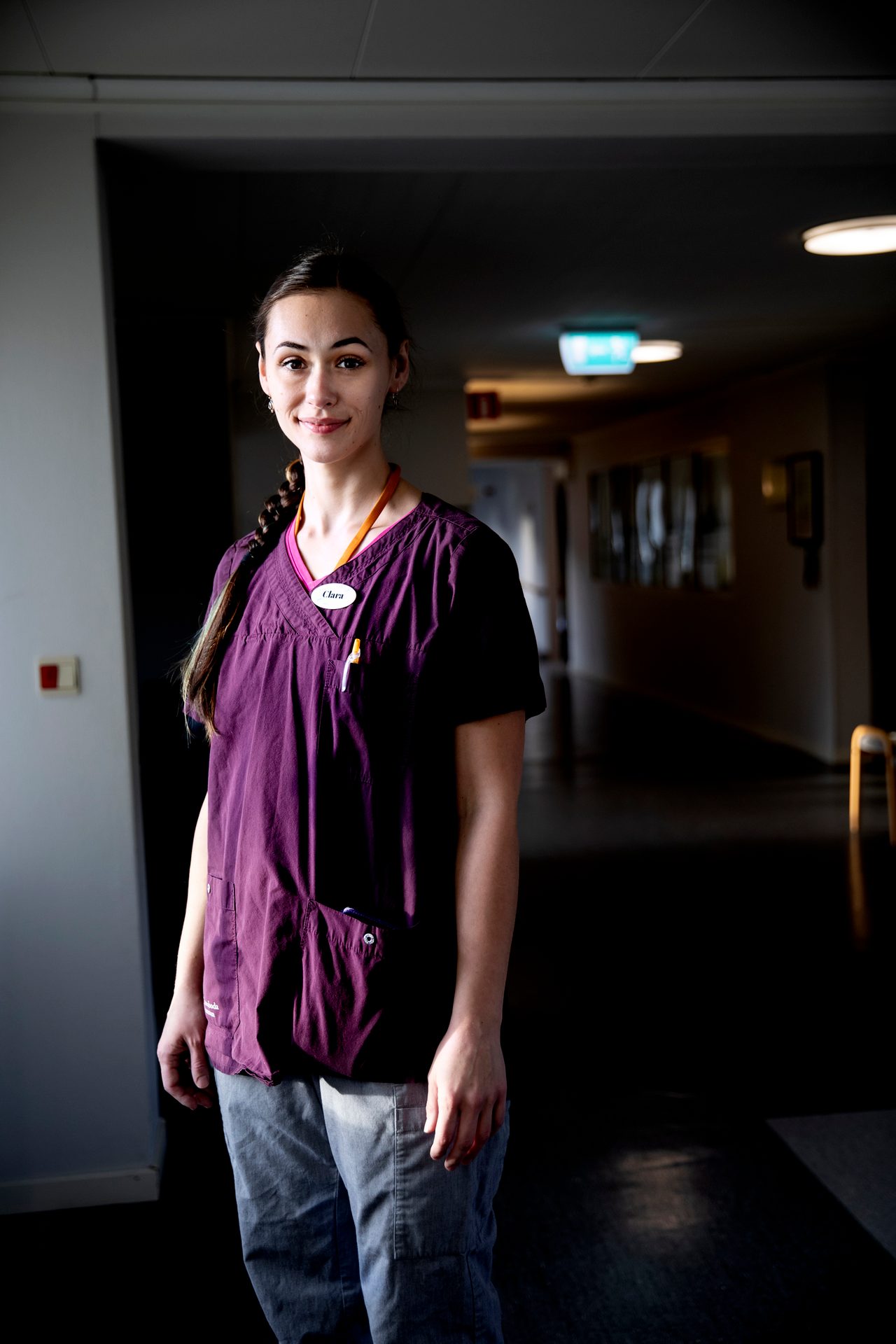 Smiling young woman in purple scrubs with a name tag stands in a hallway.