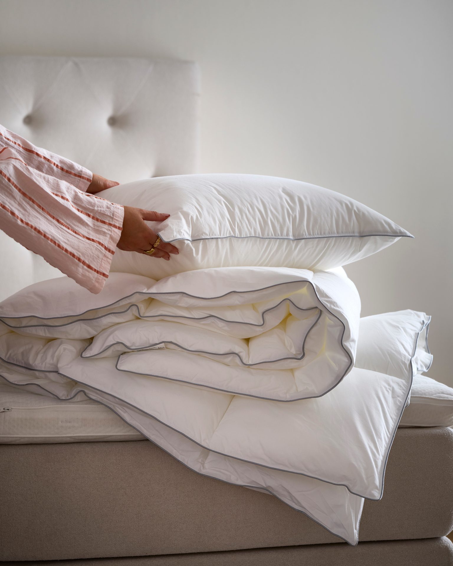Hands arrange a stack of fresh white bedding: a fluffy pillow and duvet with grey trim on a bed.