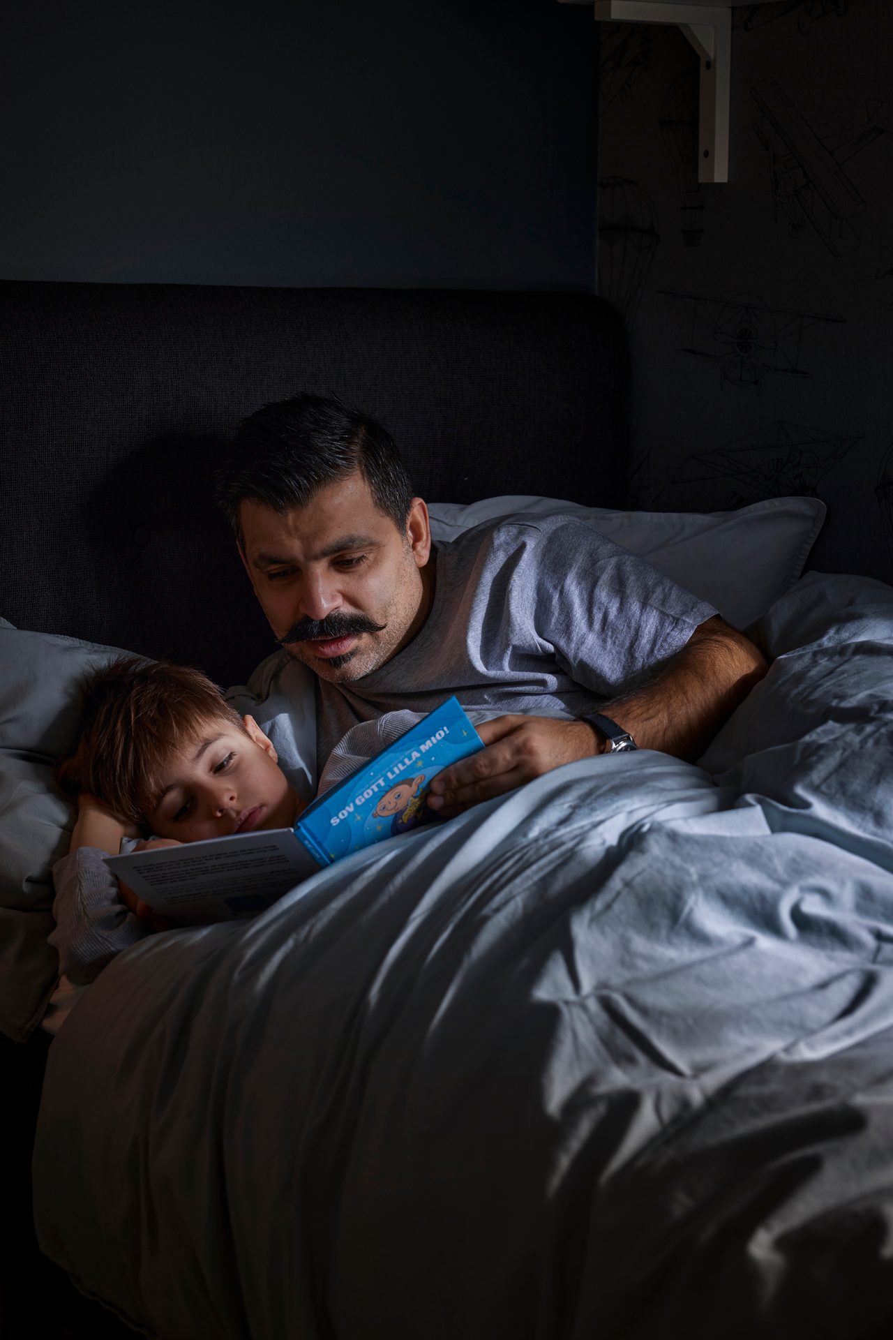 Father with a mustache reads a bedtime story to his young child, who is sleeping in bed.