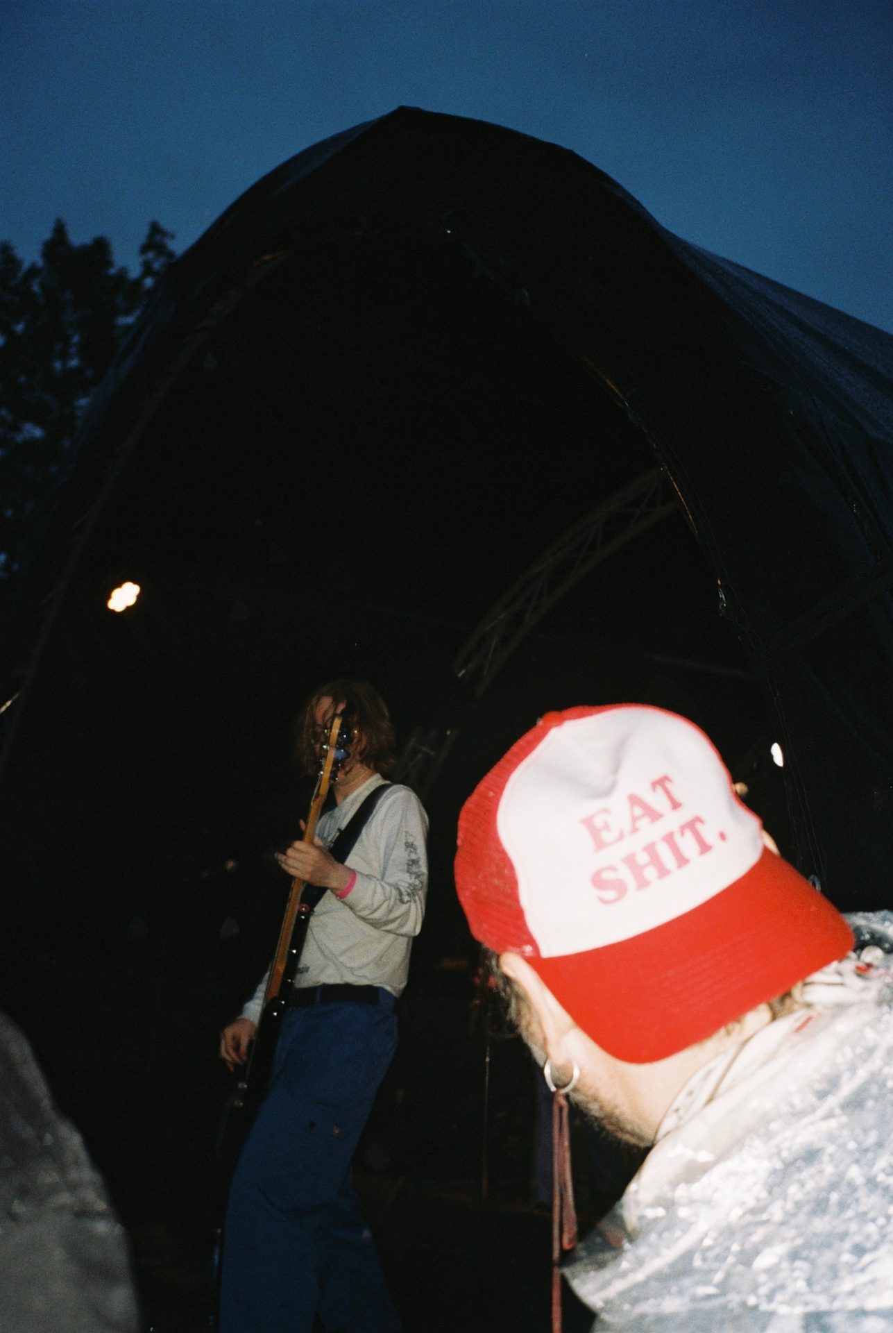 Person in "EAT SHIT" hat watches a musician playing bass guitar on a dark stage.