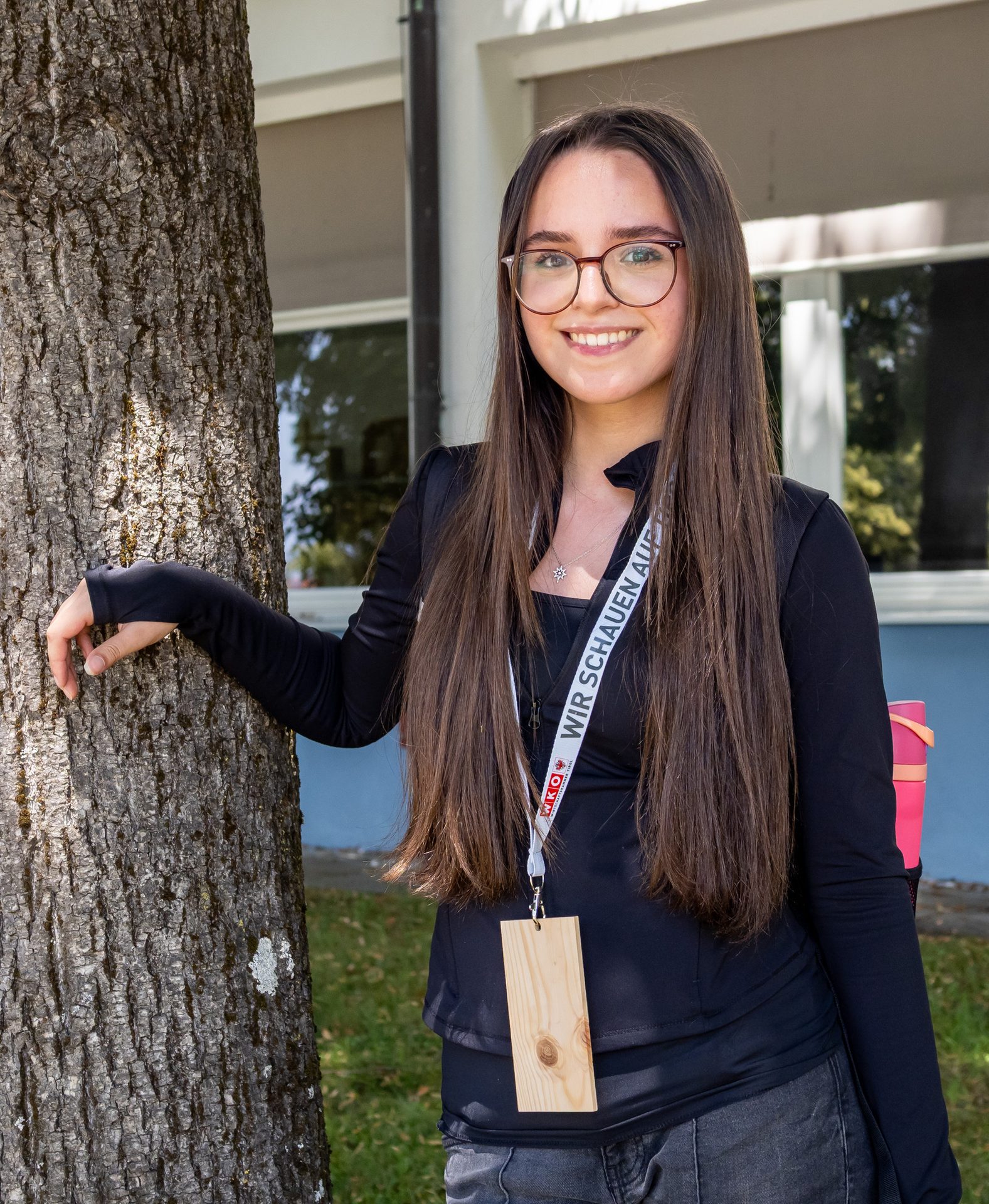 Smiling young woman with long hair and glasses, wearing a black top and lanyard, leaning against a tree.