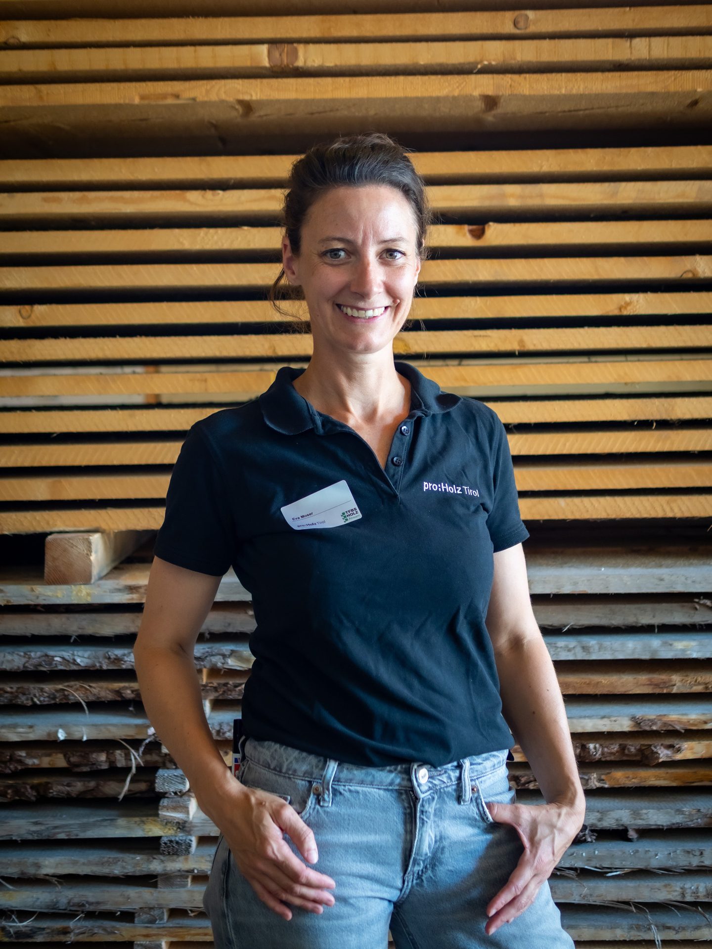 A smiling woman wearing a black polo shirt and jeans stands in front of stacks of wooden planks.