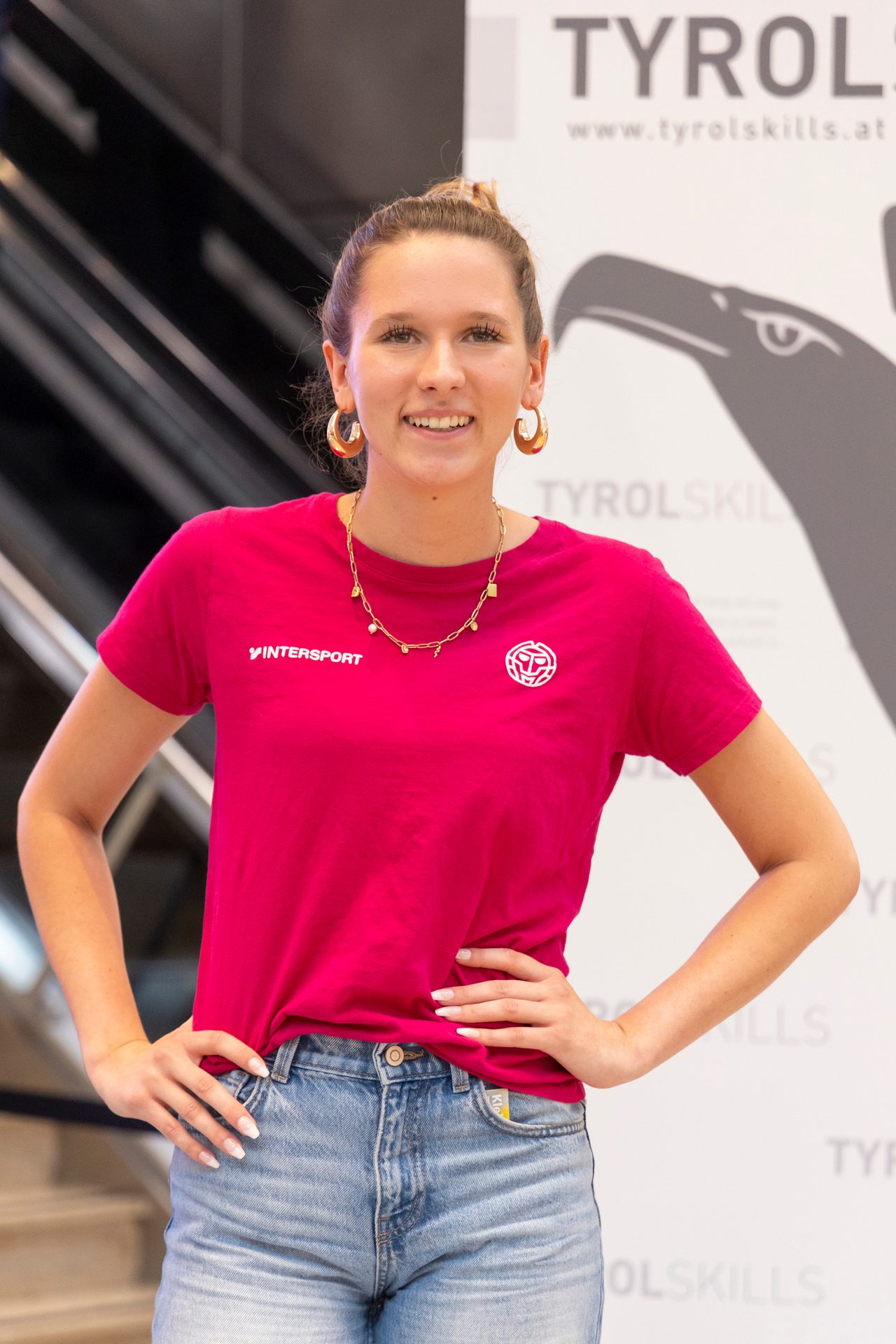 A young woman with a bun, hoop earrings, and a pink Intersport t-shirt smiles.