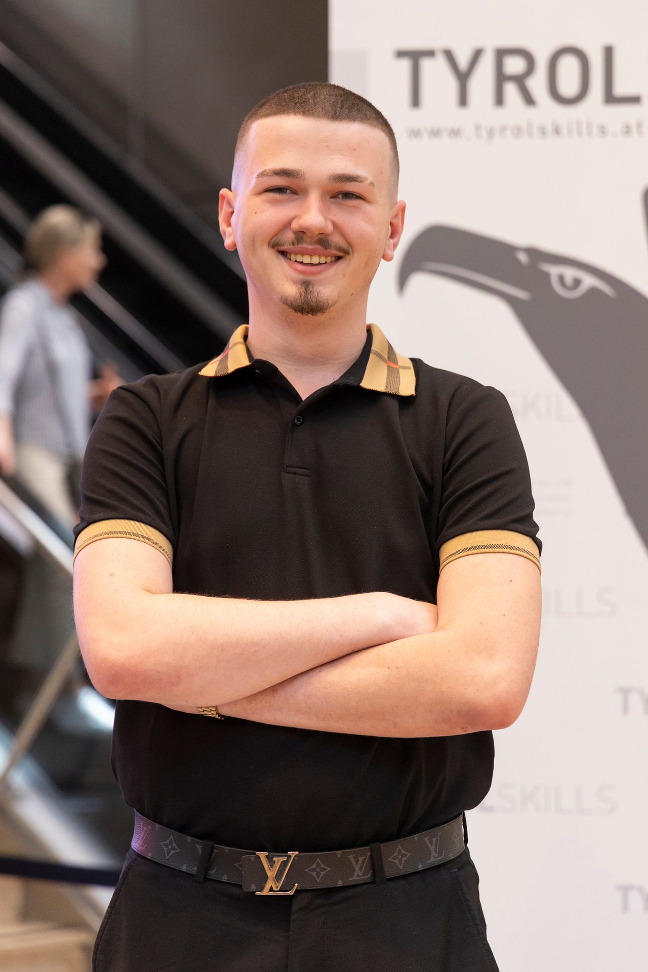 Smiling young man with buzz cut and goatee, wearing black polo with checkered trim and a designer belt.
