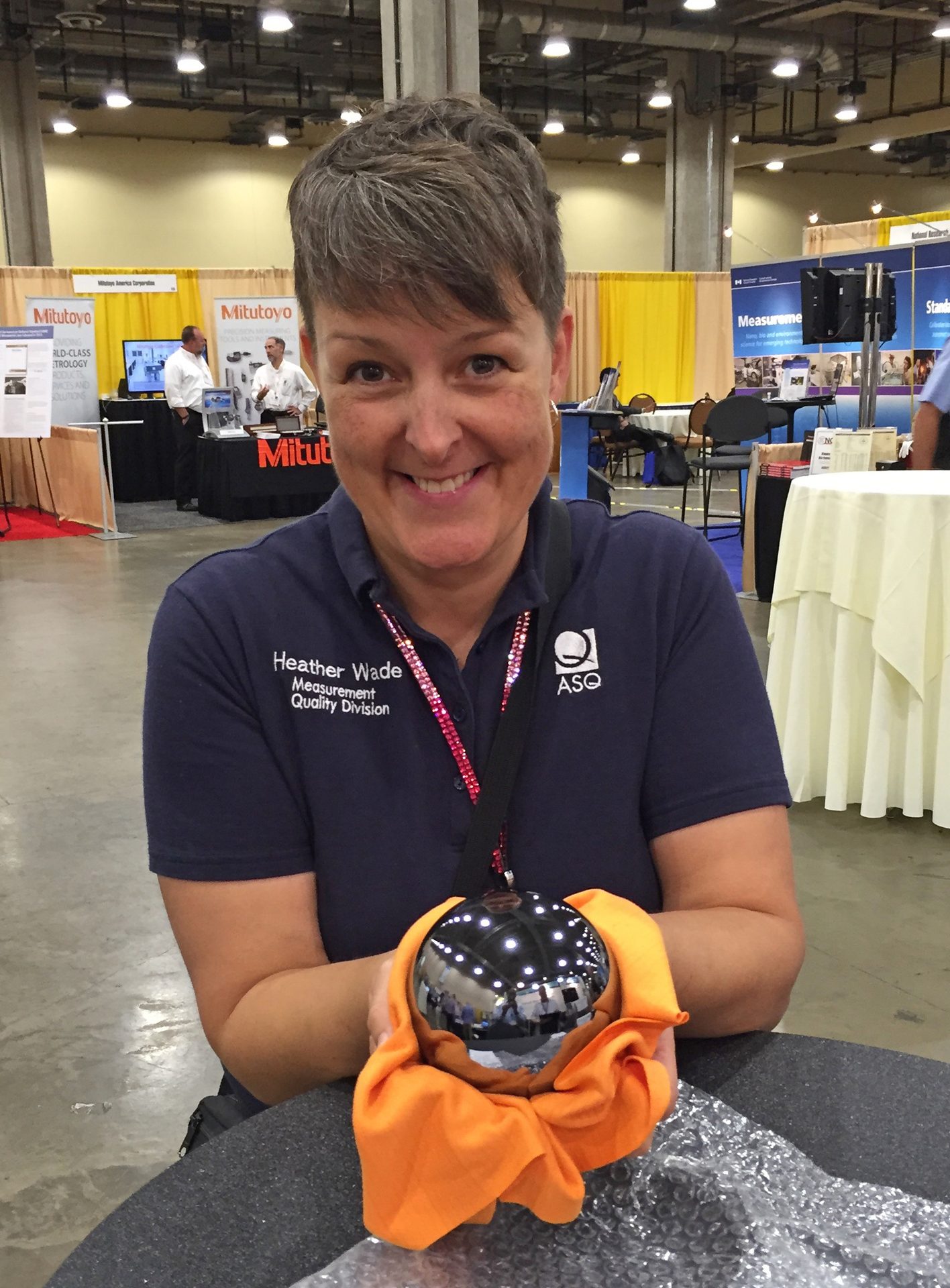 Heather Wade smiling, holding a shiny, reflective sphere at an exhibition booth with "Mitutoyo" visible.
