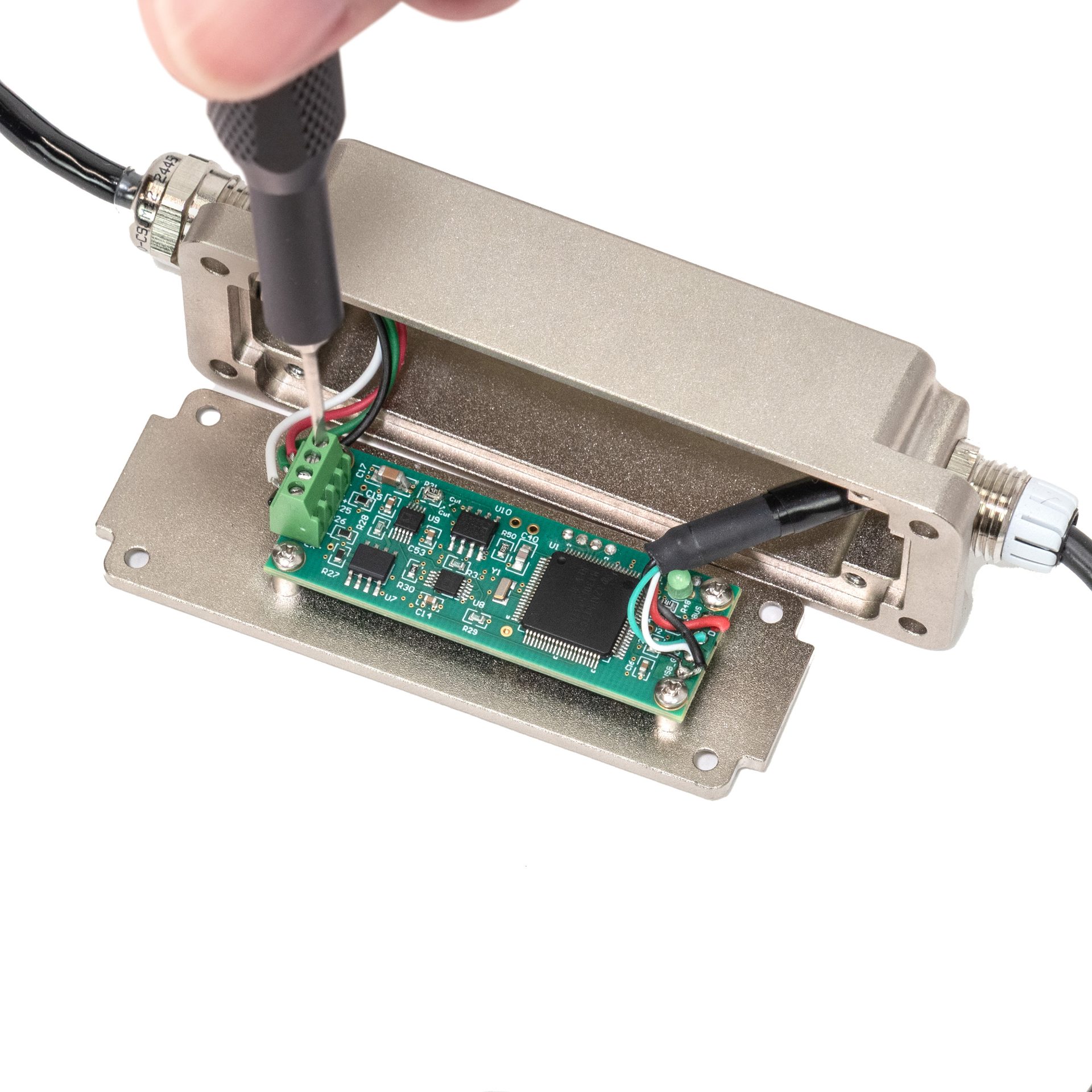 A hand uses a screwdriver to connect wires to a green circuit board inside an open metal electronics enclosure.