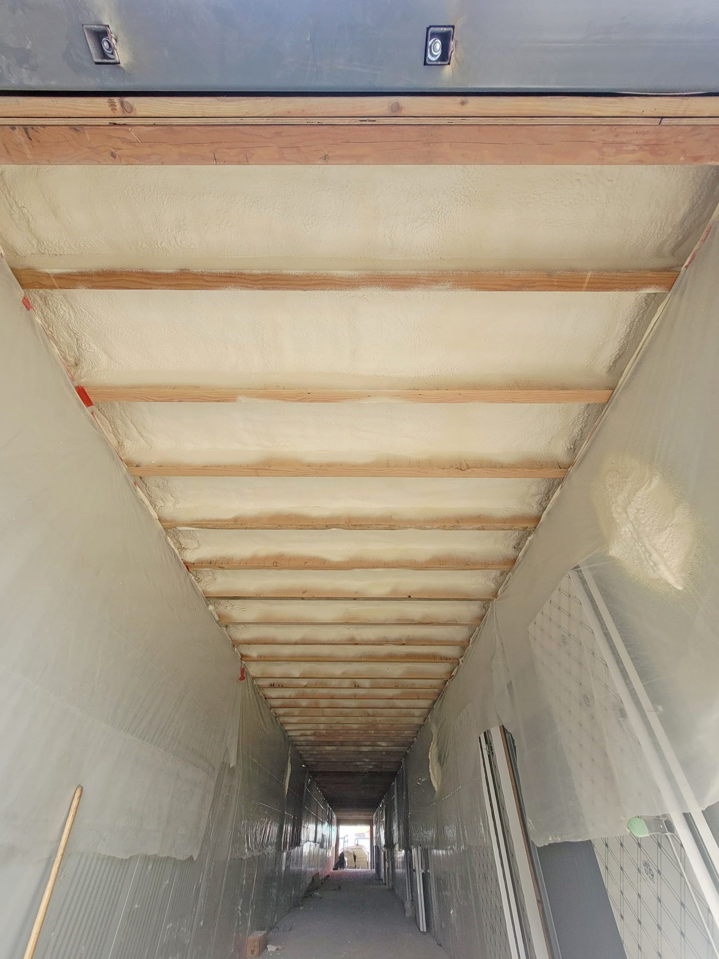 A long corridor under construction with spray foam insulation between wooden ceiling beams and covered walls.
