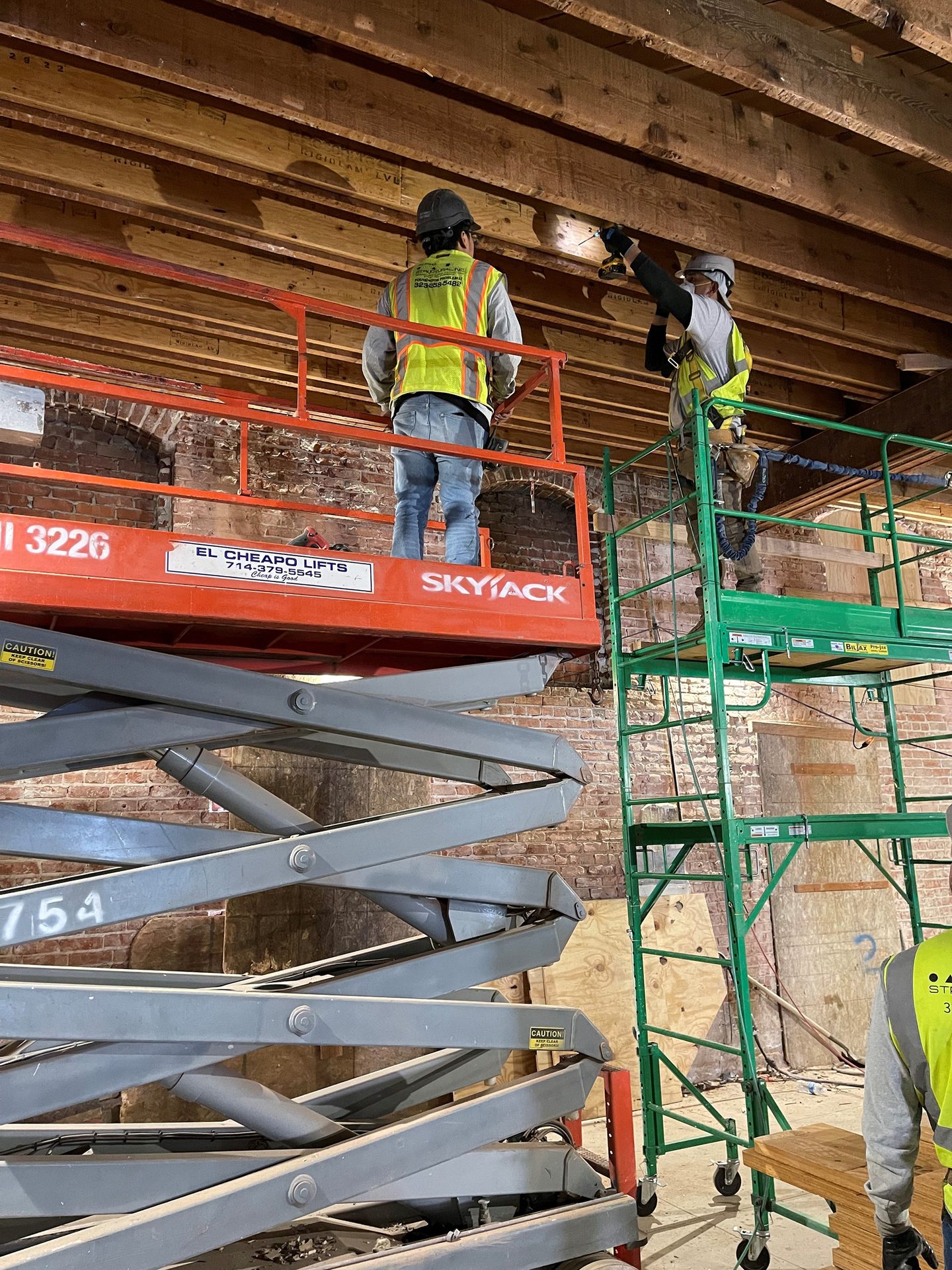 Two construction workers on lifts and scaffolding install overhead wood beams in a building.