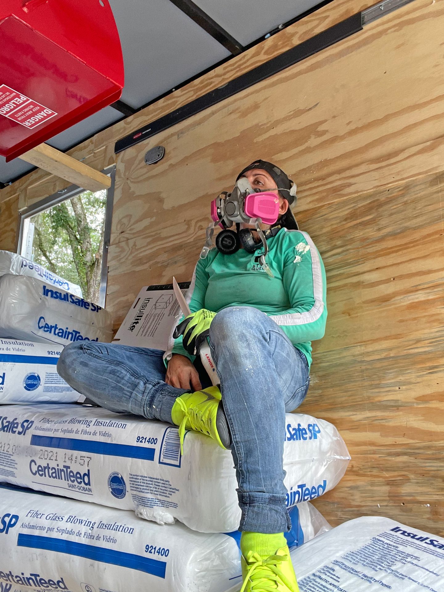 Worker in respirator mask sits on insulation bags inside a wooden trailer.