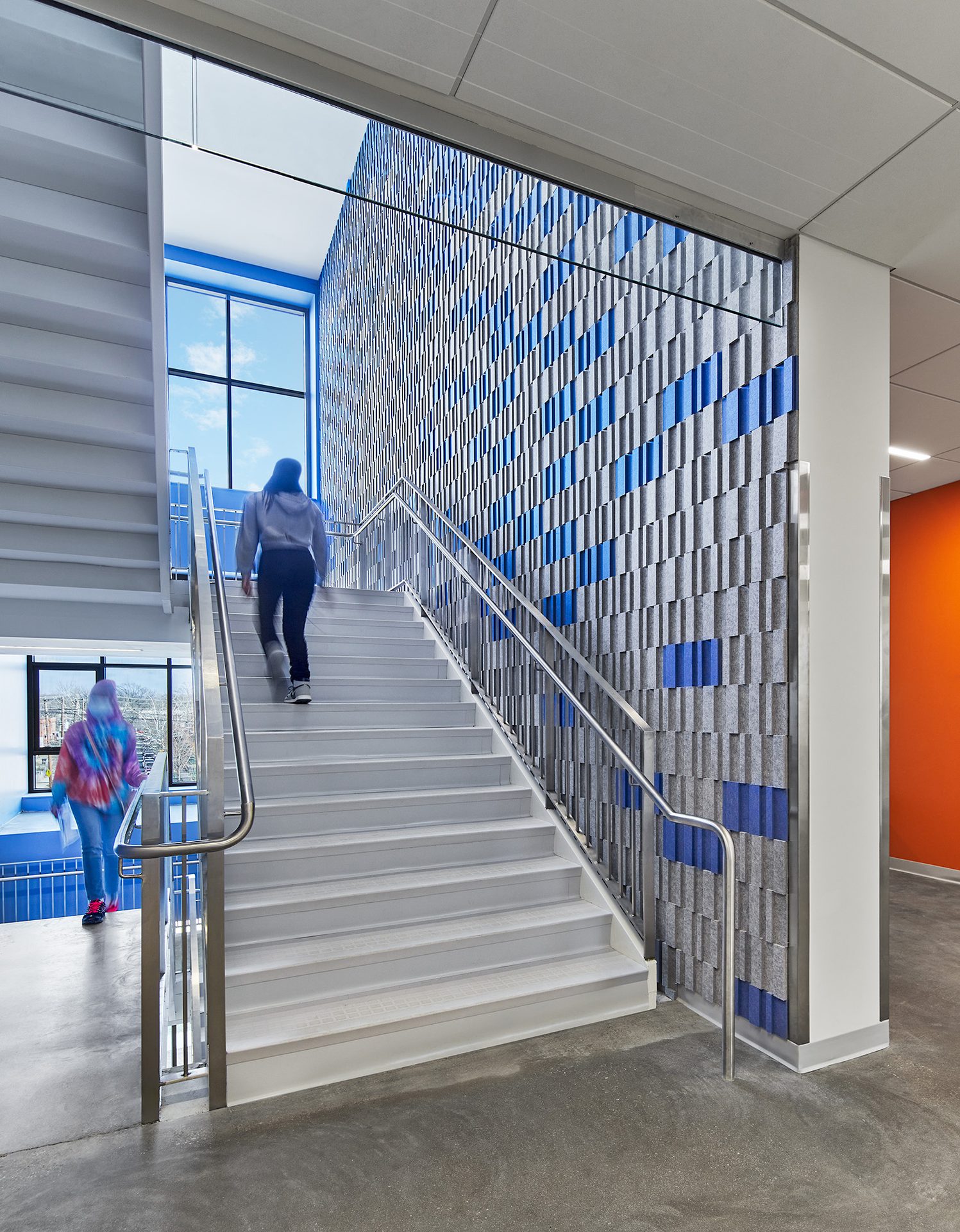Two people on white stairs next to a blue/grey textured wall, large windows, and an orange accent wall.