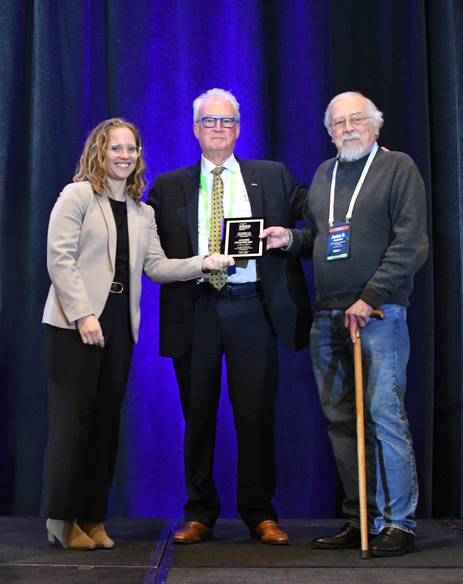 A smiling woman presents a black plaque award to two men on stage with a blue backdrop.