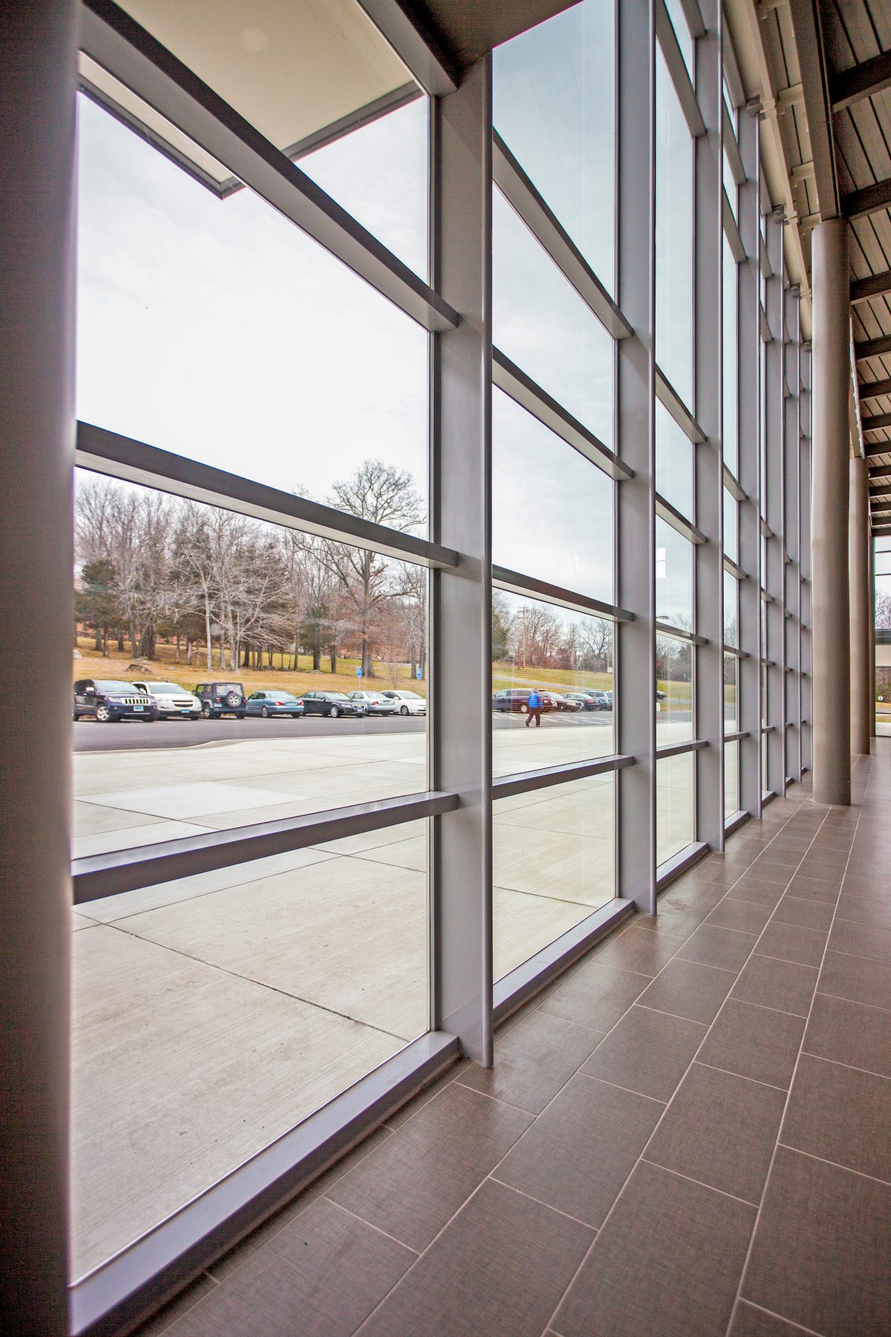 Modern building interior with a long glass wall facing a parking lot, cars, and bare trees.