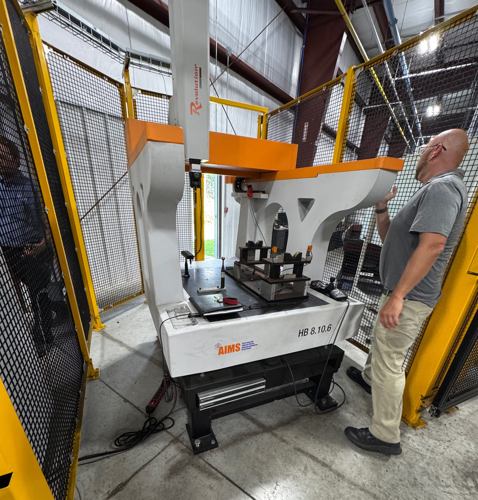 Bald man with earplugs inspecting an AIMS Revolution CMM in a factory safety cage.