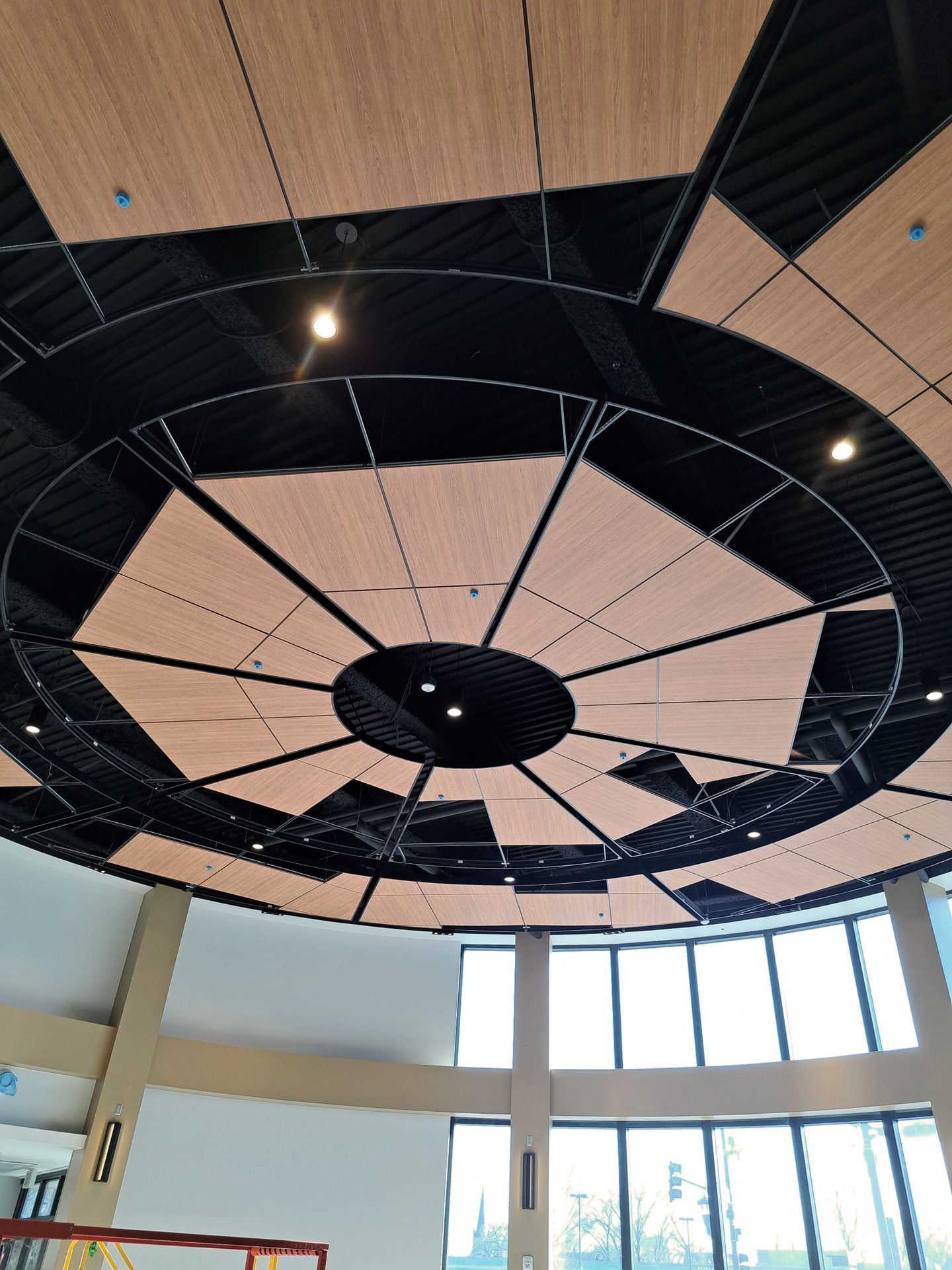 Circular ceiling with geometric wood panels and black sections, recessed lighting, above tall windows.