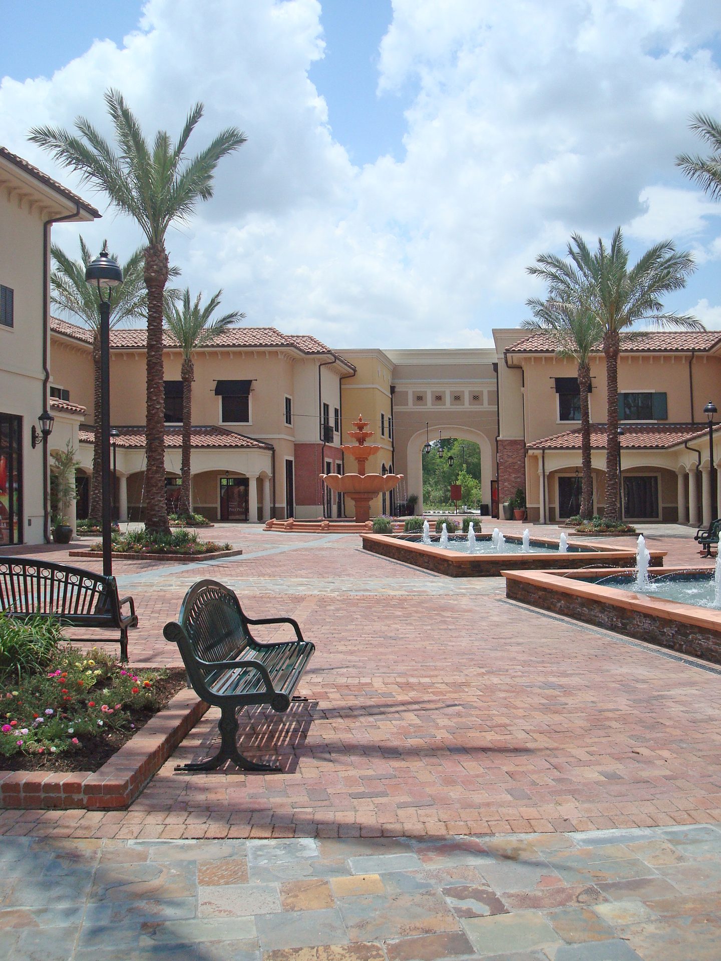 Courtyard with Mediterranean architecture, palm trees, fountains, benches, and brick pavers.