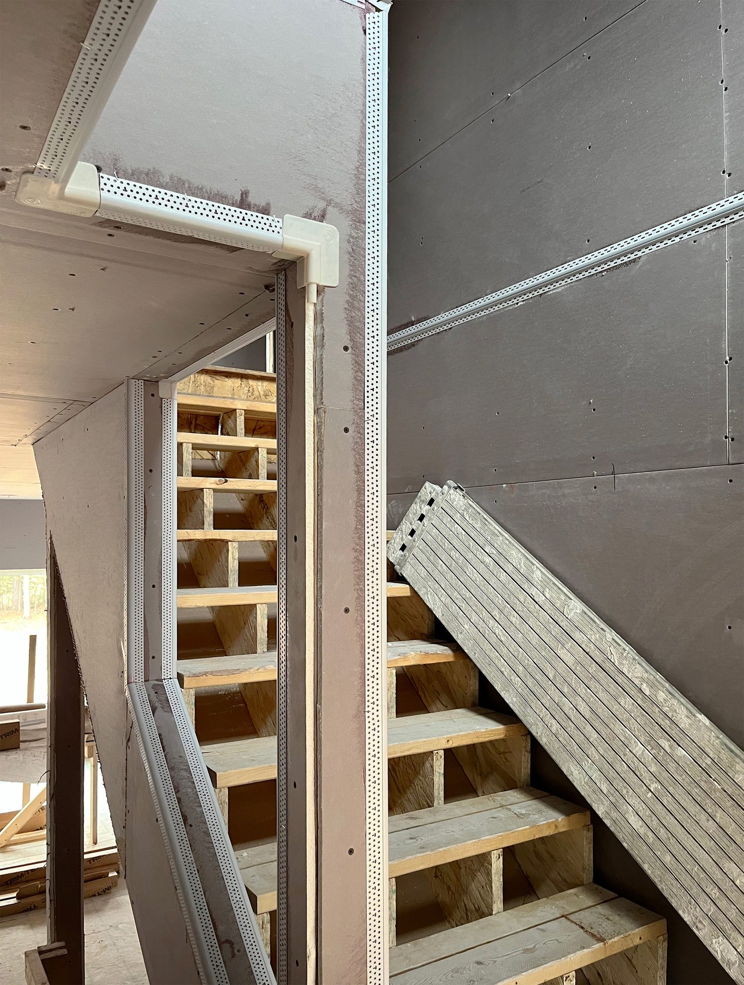 Interior construction progress with installed drywall, corner beads, and unfinished wooden stairs.