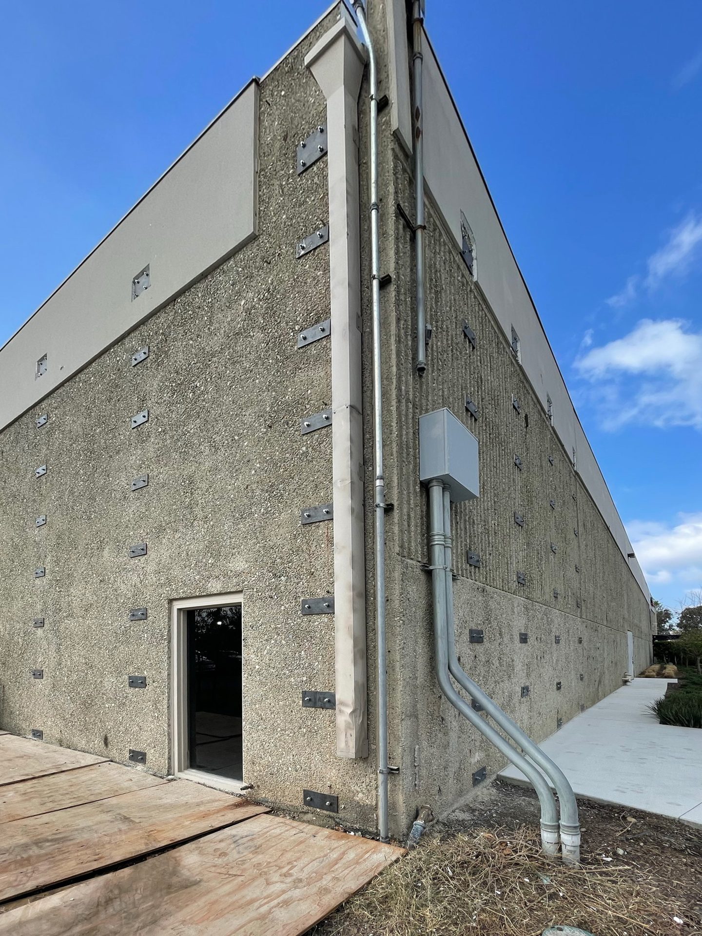 Corner view of a textured concrete building with metal plates, conduits, and a door under a blue sky.