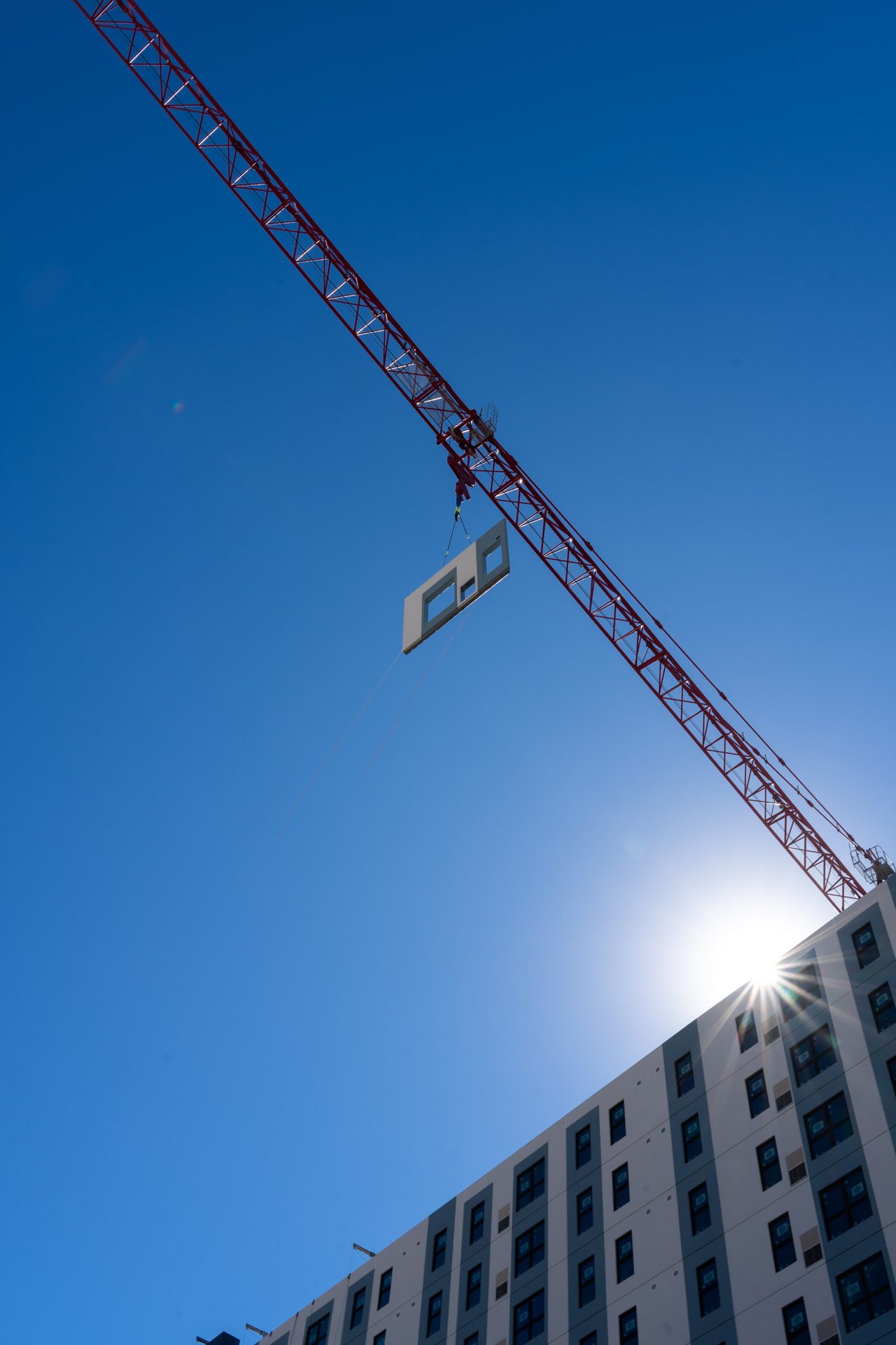Red construction crane lifting a modular wall panel against a bright blue sky, with sun and building.