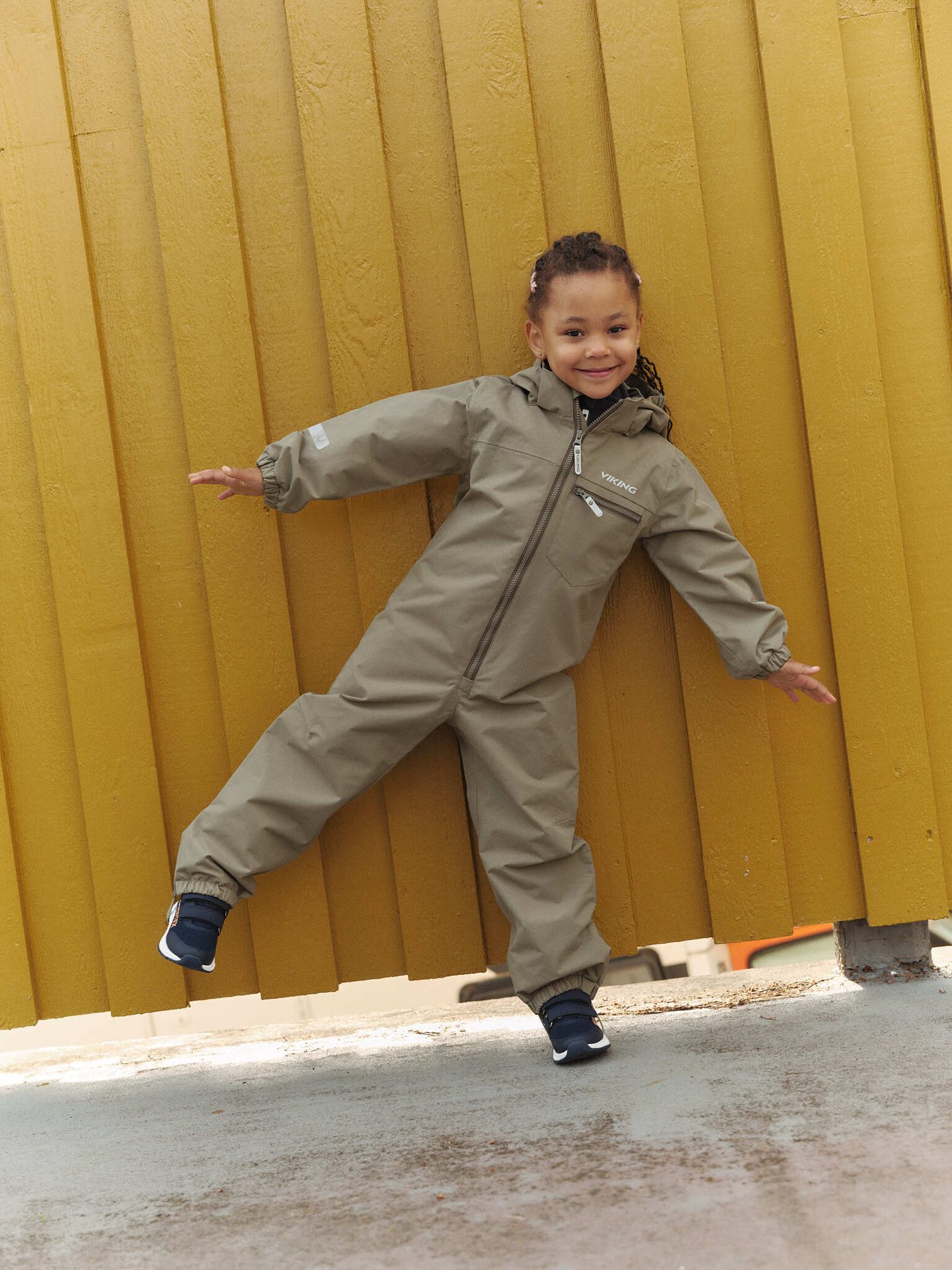 Smiling child in khaki jumpsuit balancing against a yellow wall.