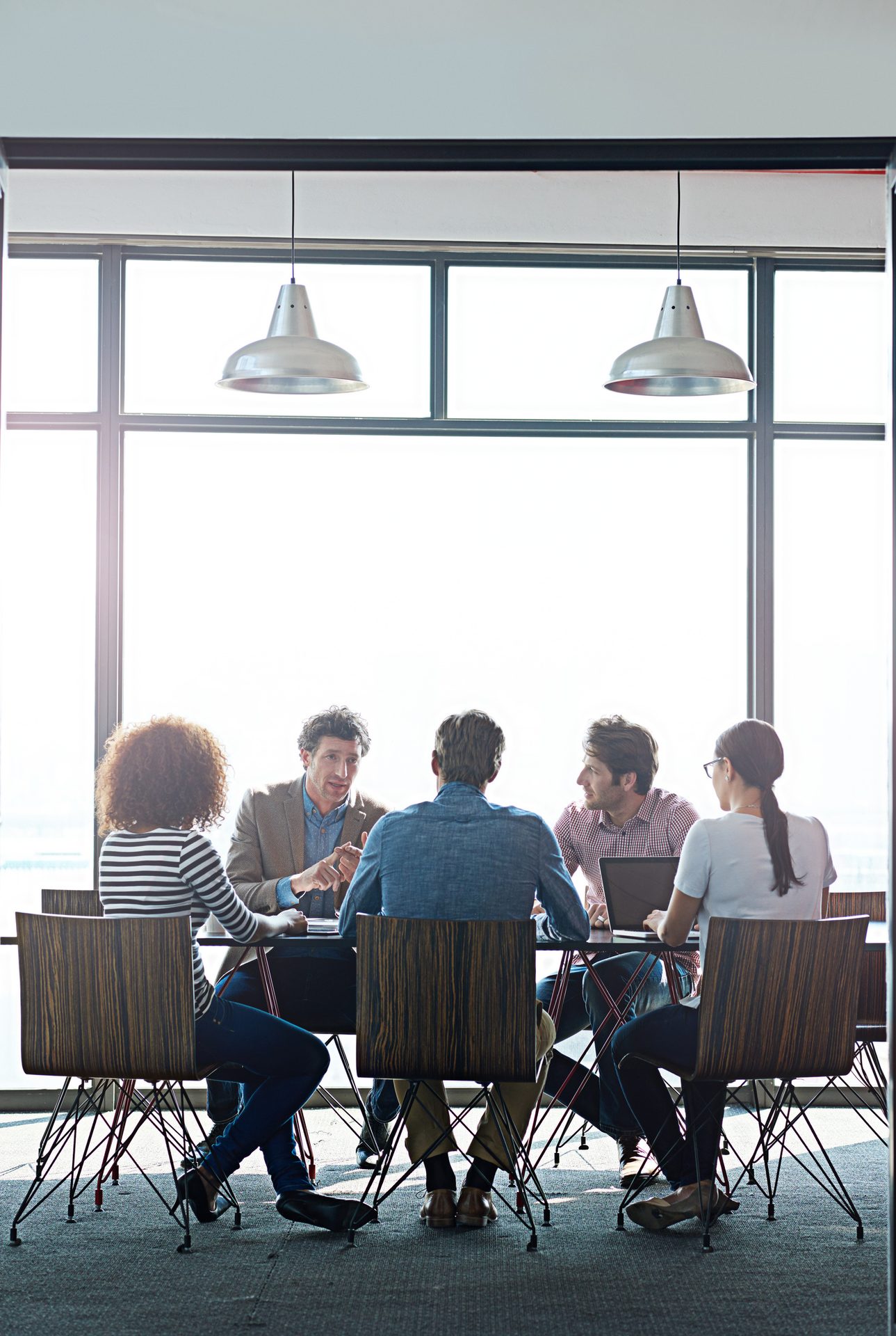 Five professionals discuss ideas around a conference table with large windows in a modern office.