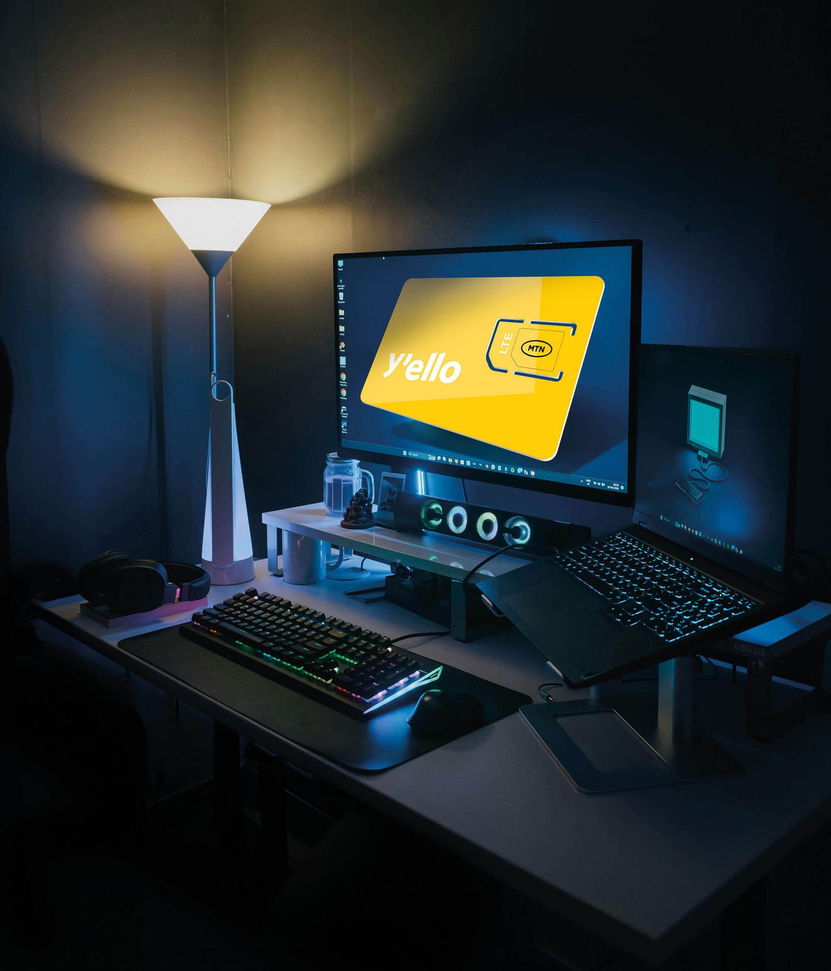 Dimly lit desk setup with an MTN SIM card on a monitor, RGB keyboard, laptop, and standing lamp.