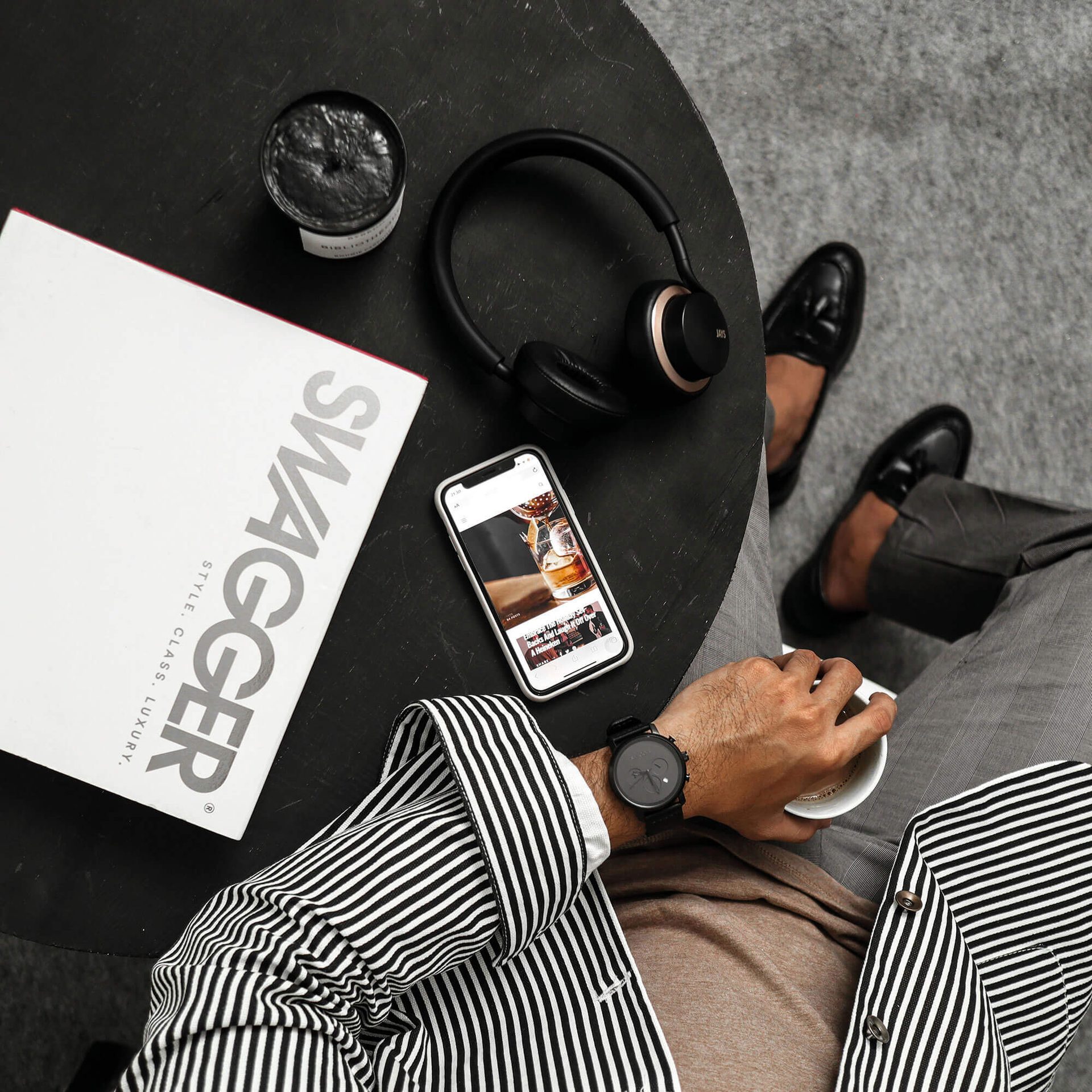 Overhead view of a person holding coffee with a phone, headphones, and "Swagger" magazine on a table.