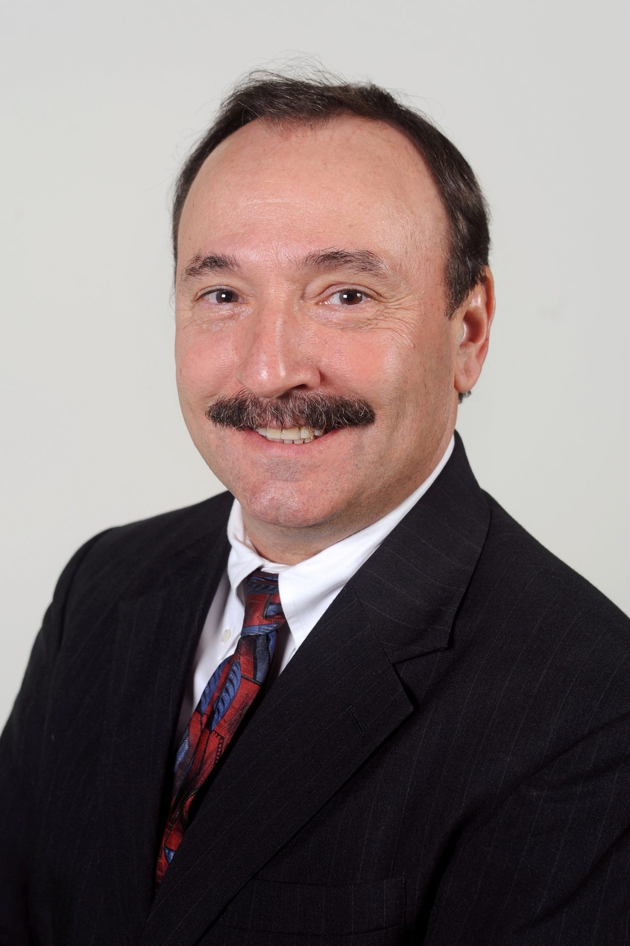Smiling man with a mustache, wearing a dark pinstripe suit and patterned tie.