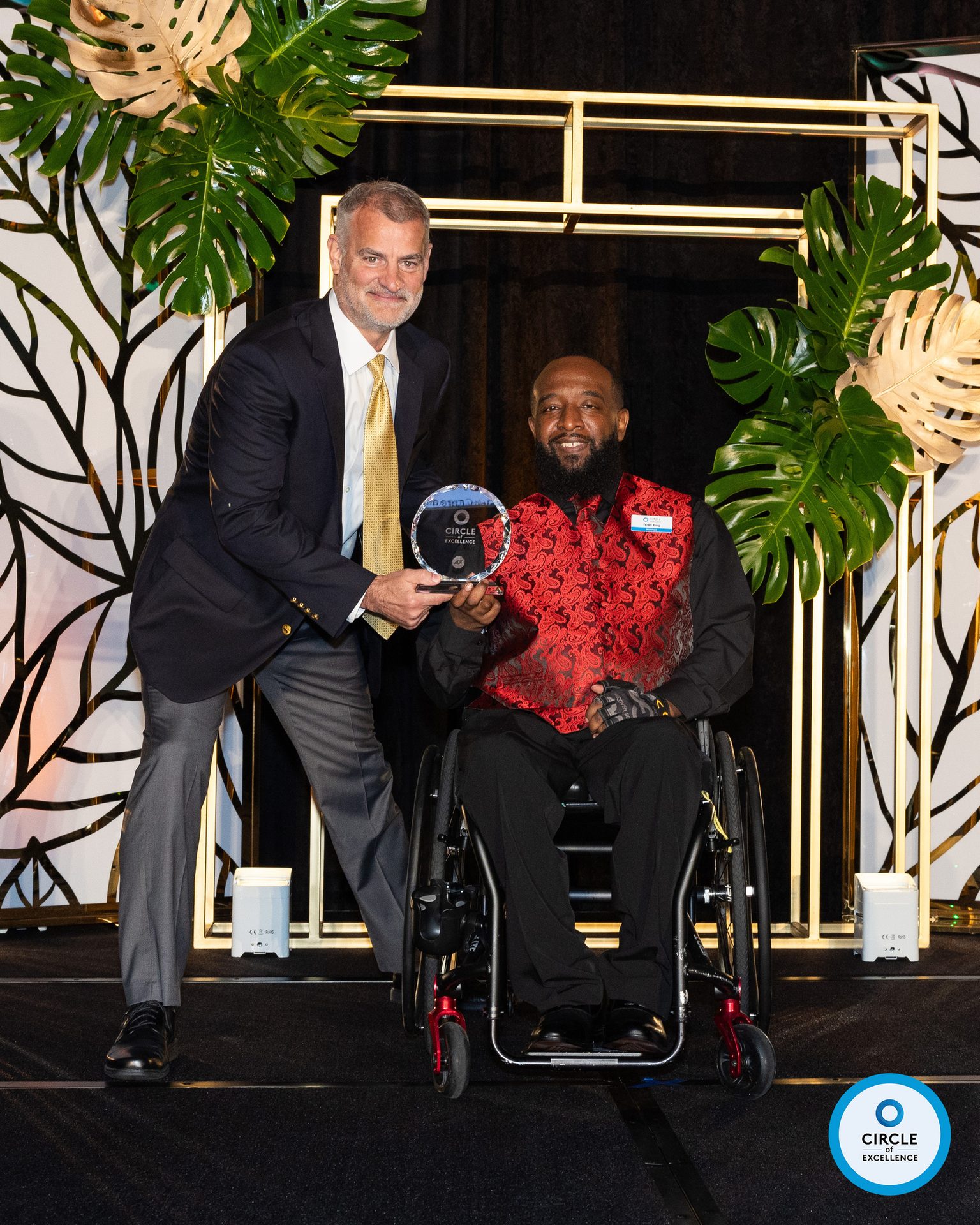 Man in suit presents Circle of Excellence award to smiling man in wheelchair.