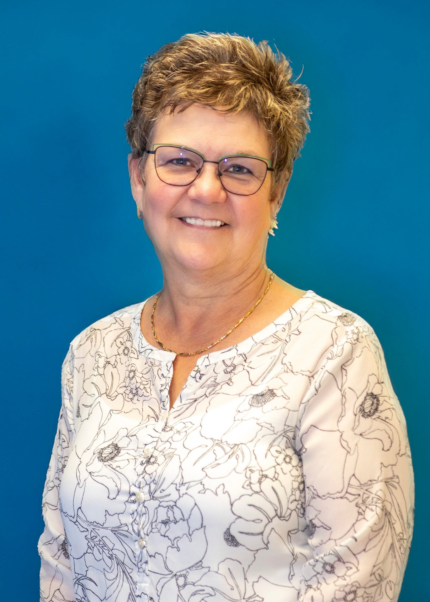 Smiling woman with short brown hair and glasses in a white floral shirt on a blue background.