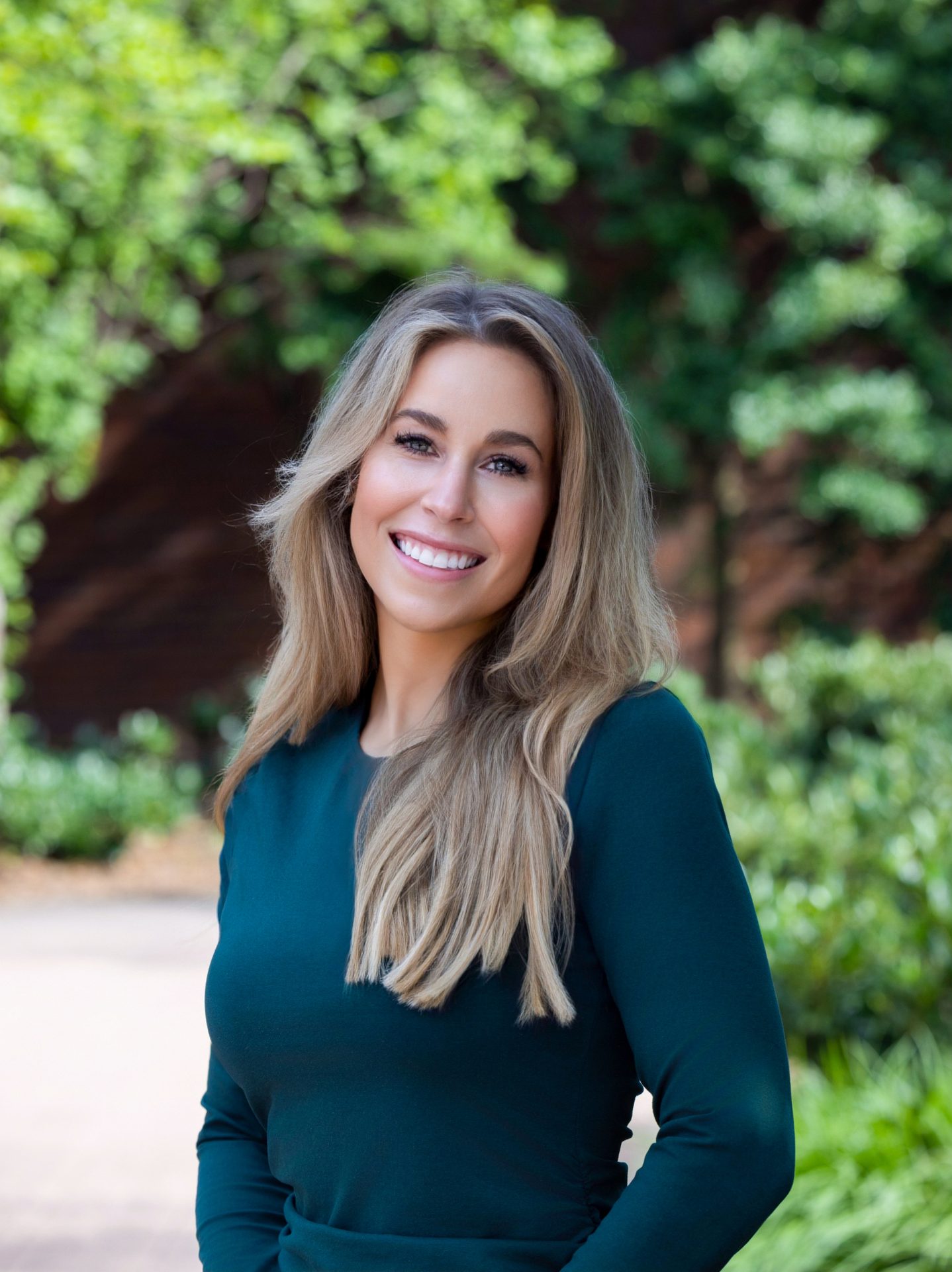 A smiling woman with long blonde hair in a dark green top, against a blurry green background.