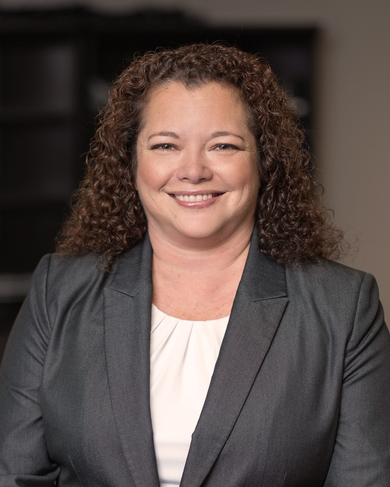 Smiling woman with curly brown hair in a grey suit, professional headshot.