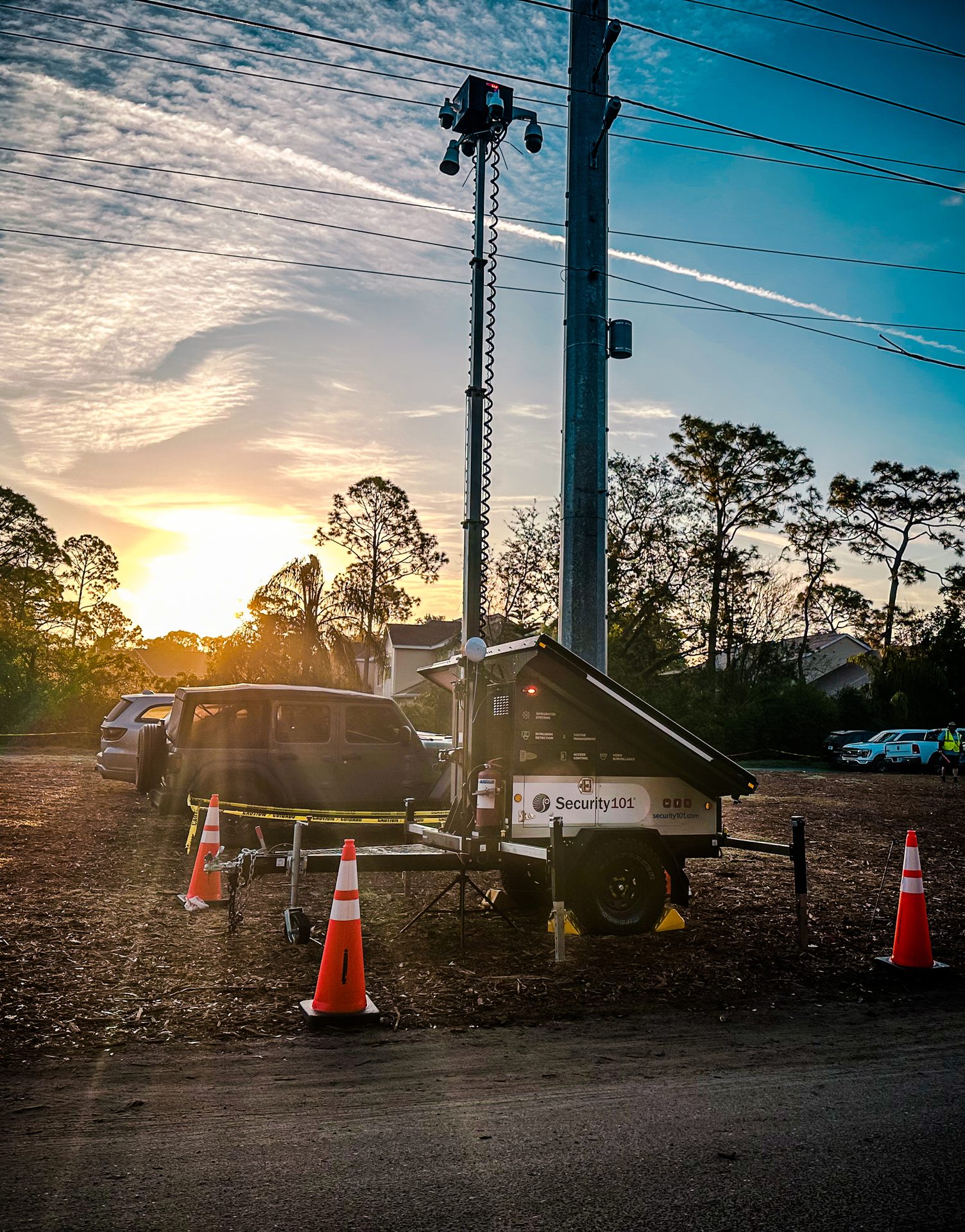 Mobile security surveillance trailer with a tall camera mast next to a utility pole at sunset.