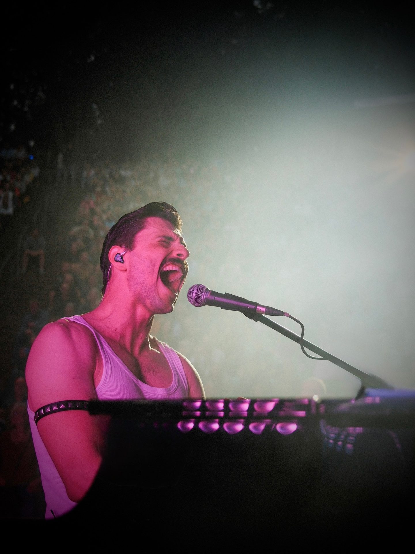 A performer resembling Freddie Mercury singing passionately into a microphone on stage, under purple light.