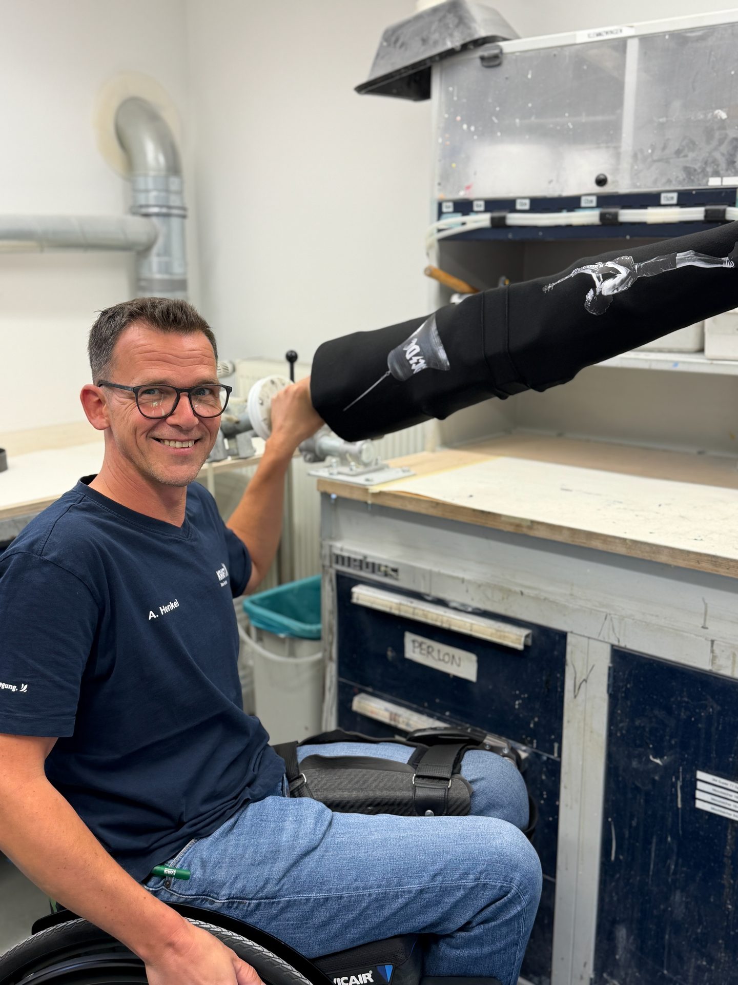 Man in a wheelchair in a workshop, smiling while working on a black prosthetic device.