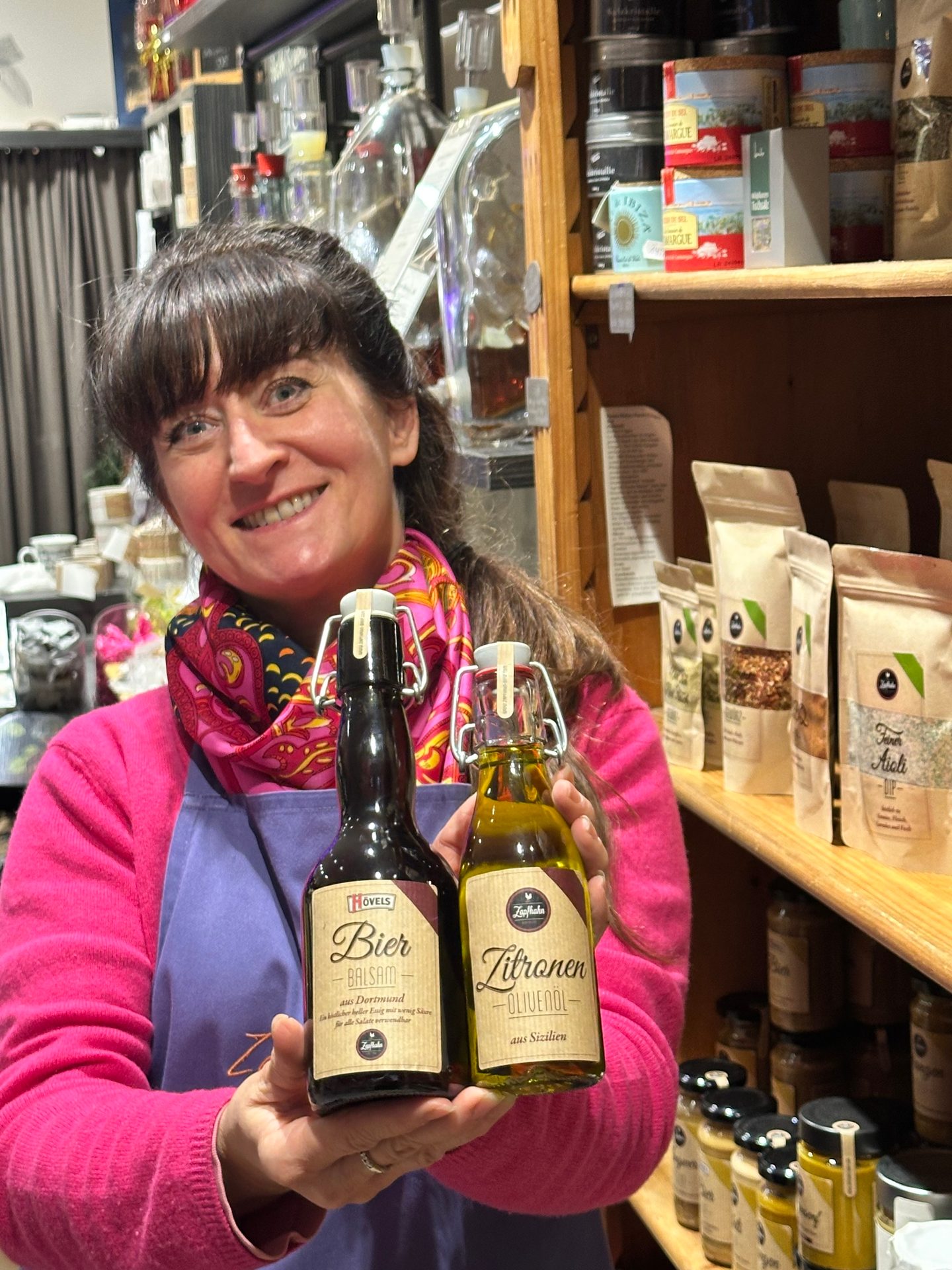 A smiling woman in a shop holds two small bottles: "Bier Balsam" and "Zitronen Olivenöl".