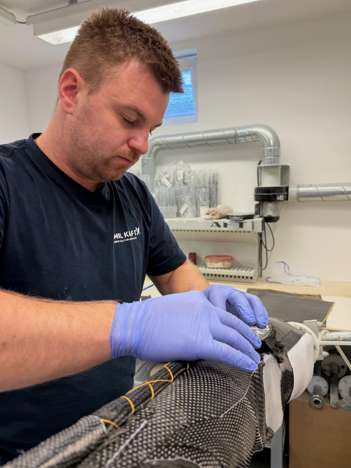 Man in blue gloves precisely working on a carbon fiber component, showcasing craftsmanship in a workshop.