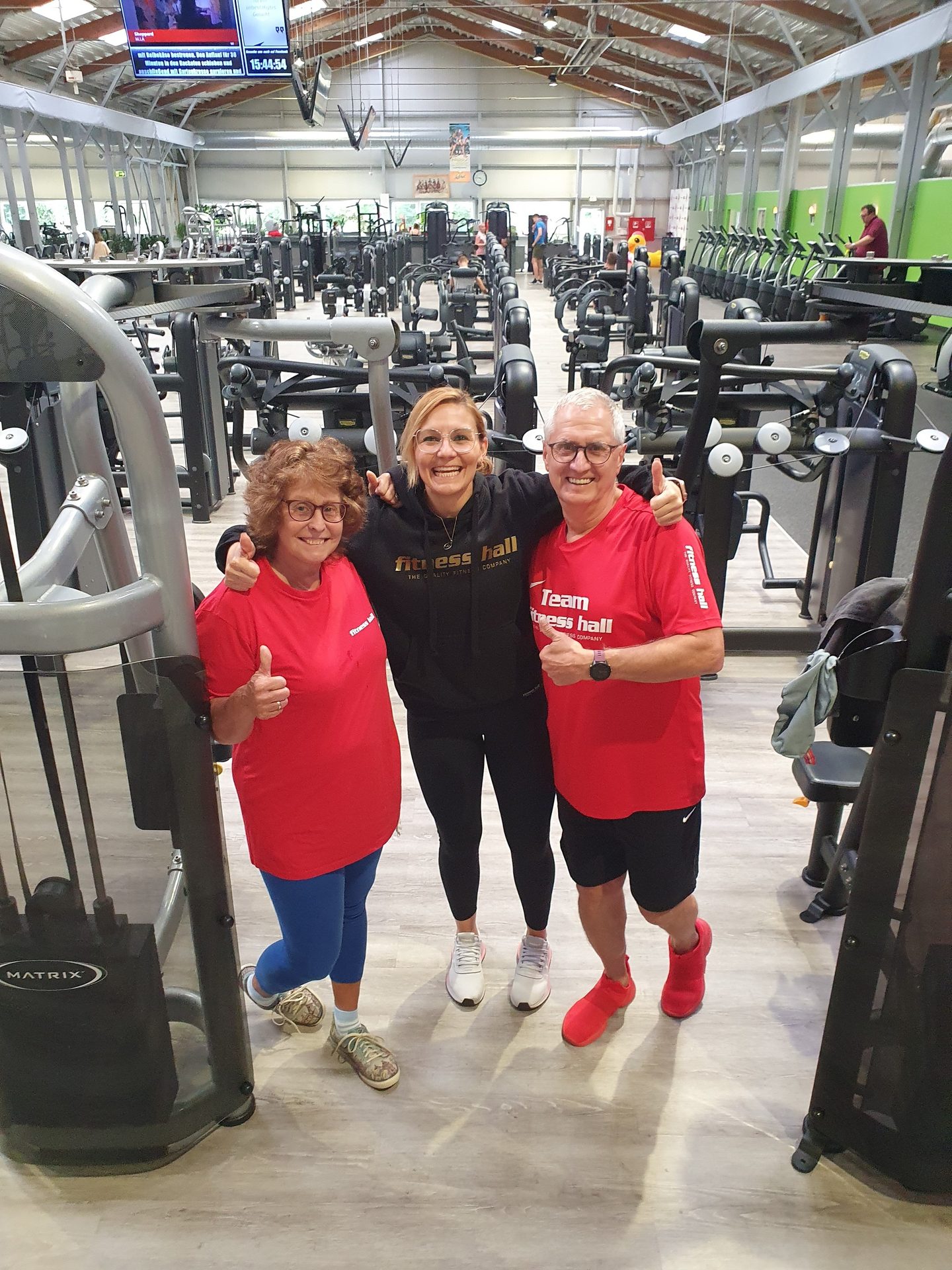 Three smiling people in "fitness hall" gear give thumbs up in a gym.
